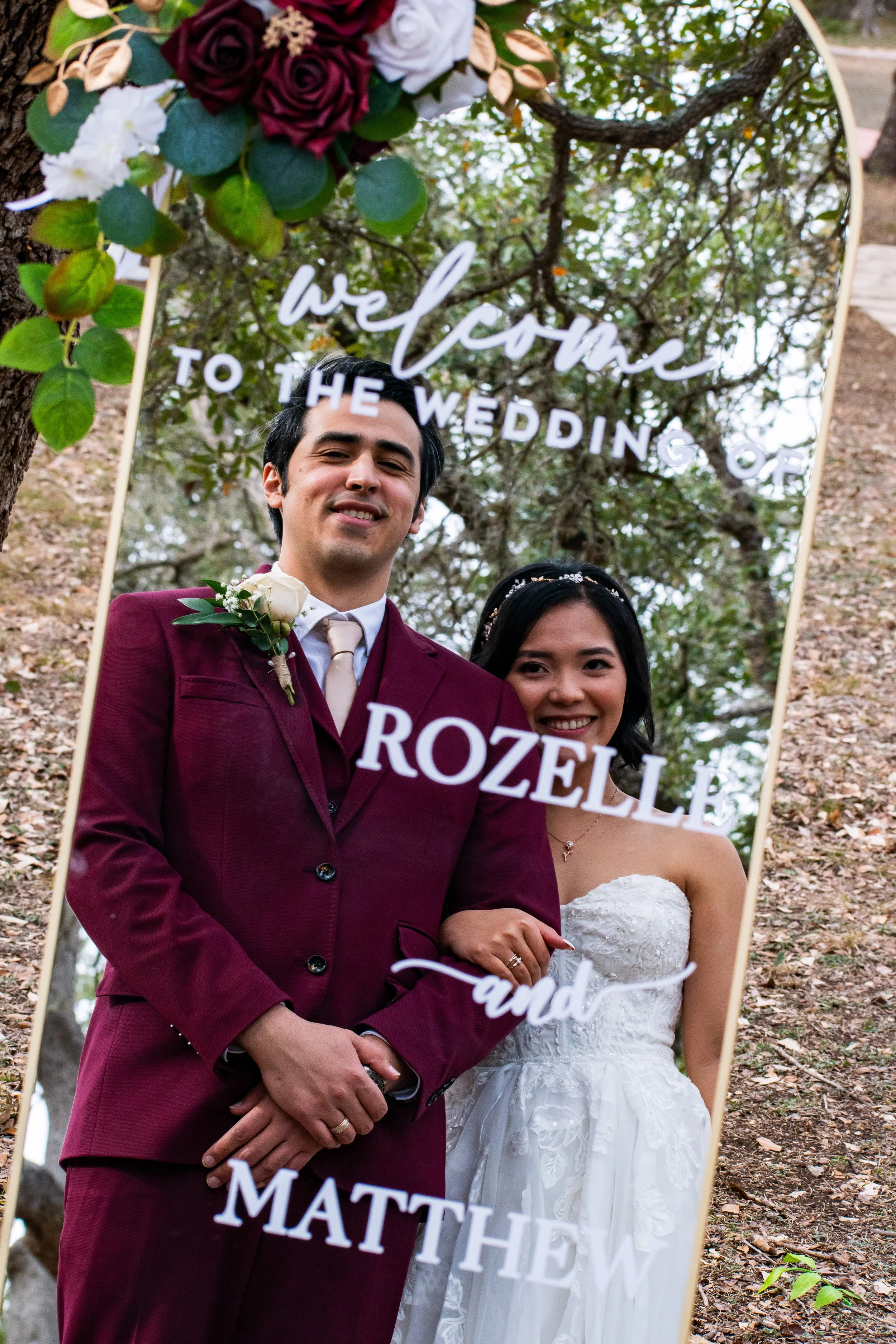 A newlywed couple stands outdoors under a tree, smiling at the camera, with a large mirror behind them reflecting the natural setting. The groom wears a burgundy suit with a boutonniere, and the bride wears a white lace wedding dress and a headband. 