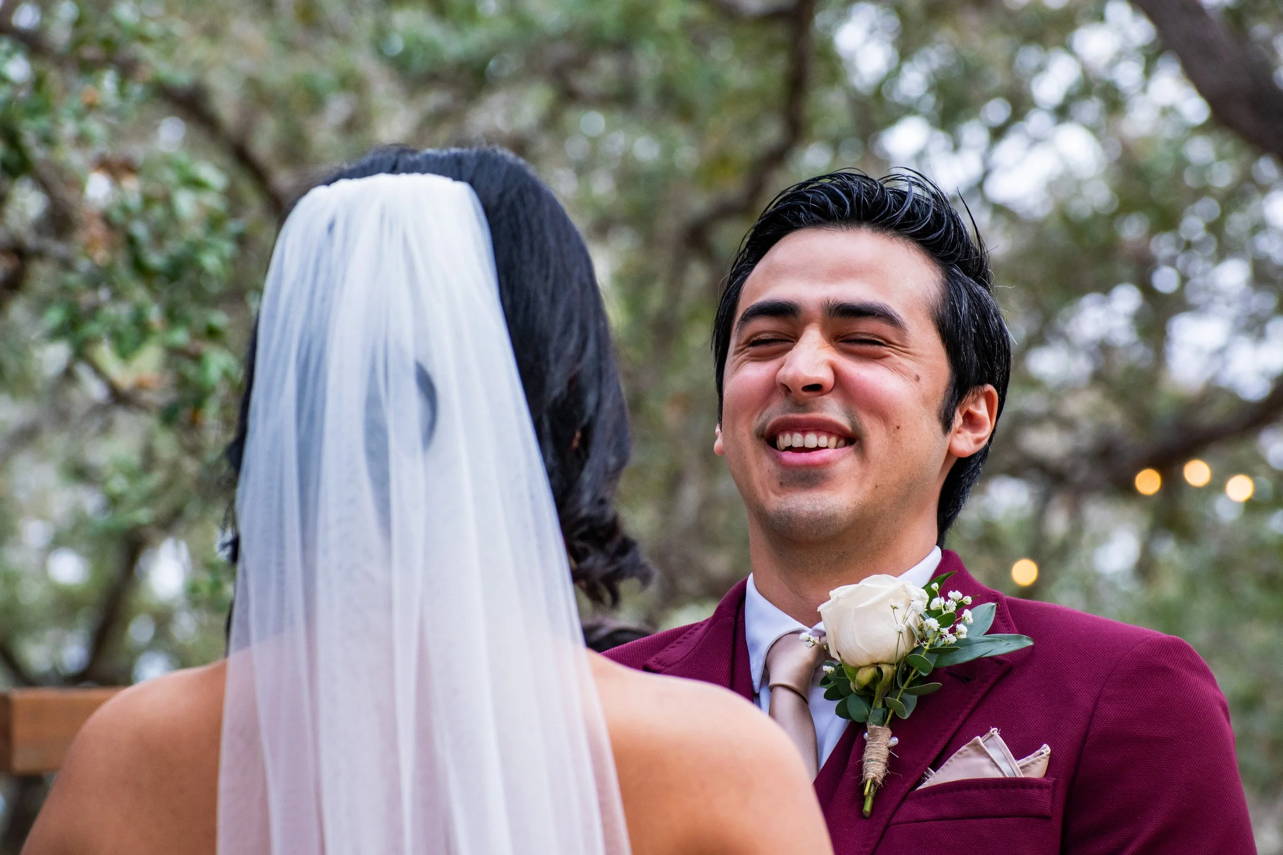 A man in a burgundy suit smiling during a wedding ceremony outdoors. The background shows trees and a blurred sky.