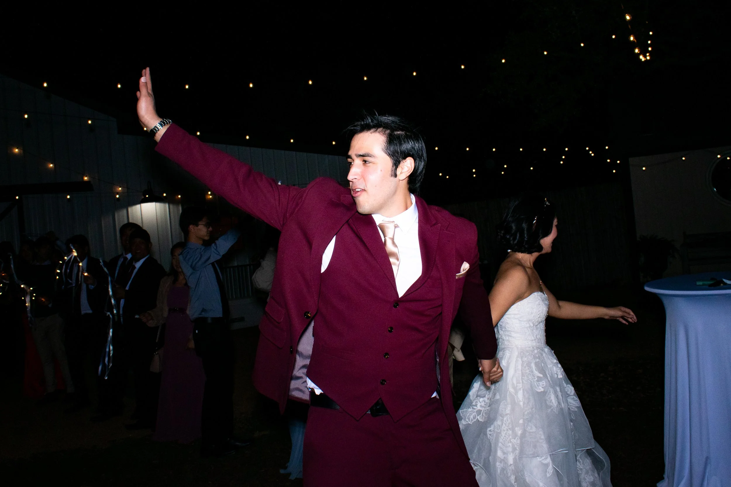 A young man in a burgundy suit dancing at a wedding reception with a woman in a white wedding dress.