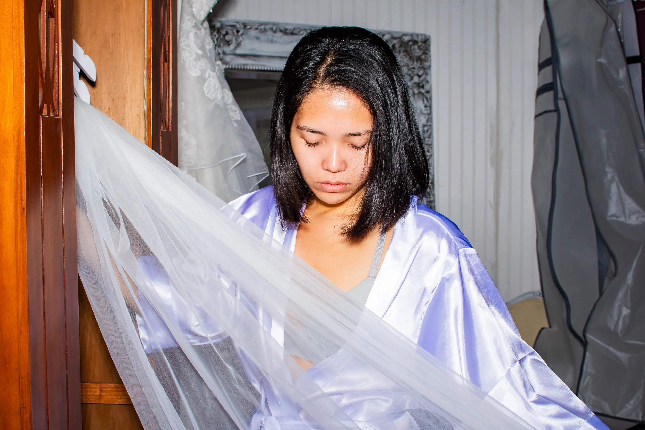 A woman with long black hair wearing a blue and white satin robe or dress, standing in front of a wooden wall or door, with her eyes closed and her head slightly bowed, surrounded by sheer white fabric or curtains.