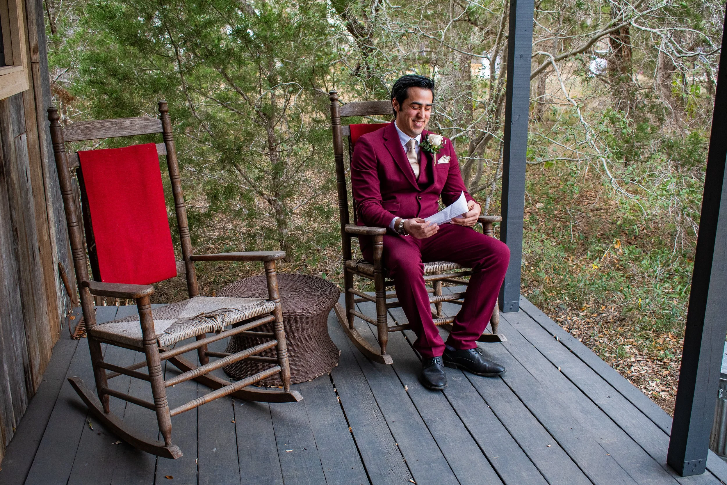 A man in a maroon suit sitting on a wooden chair on a porch, holding a letter and smiling, with trees and foliage in the background.