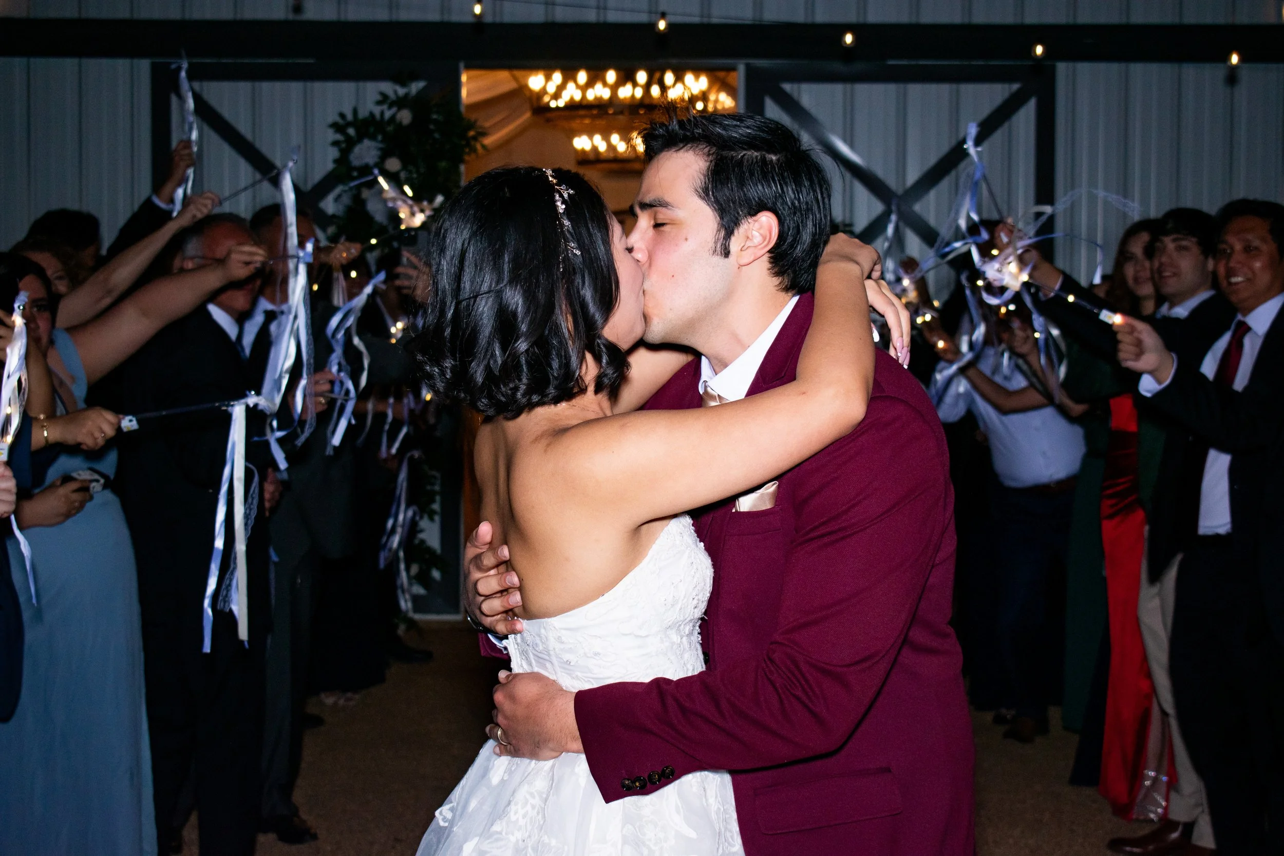 A couple sharing a kiss while dancing at a wedding reception, surrounded by guests celebrating with streamers and fairy lights.