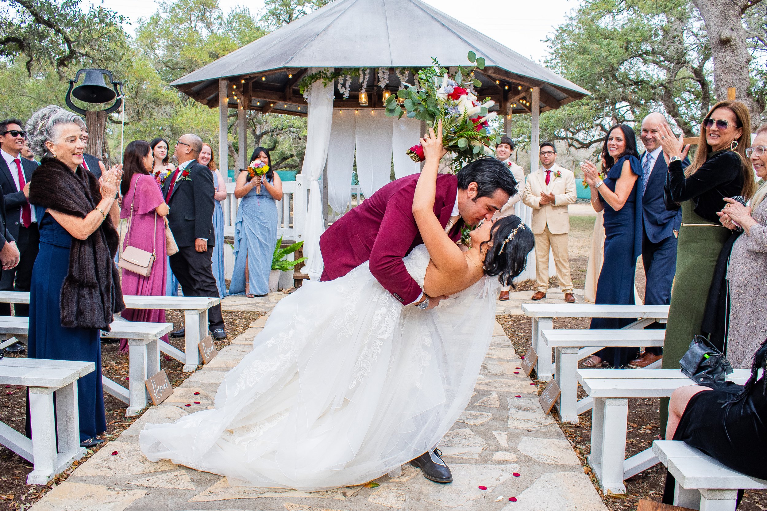 A bride and groom kiss during their outdoor wedding ceremony, with guests smiling and clapping around them, under a gazebo decorated with flowers and string lights.
