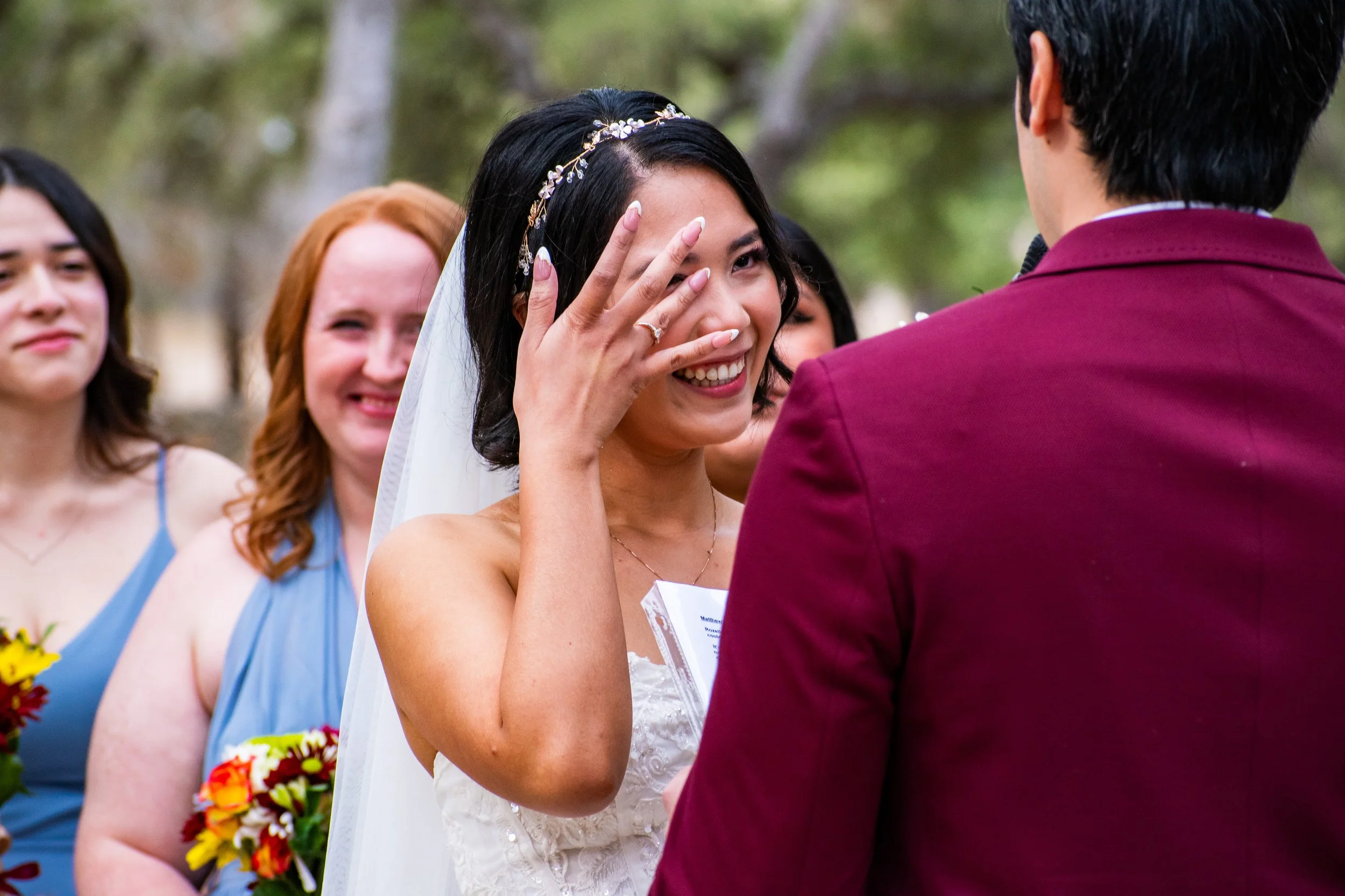 A bride with dark hair and a floral headband is smiling and wiping tears during her wedding ceremony while holding her hand near her face, in front of a groom in a maroon jacket, surrounded by bridesmaids in colorful dresses and holding bouquets.