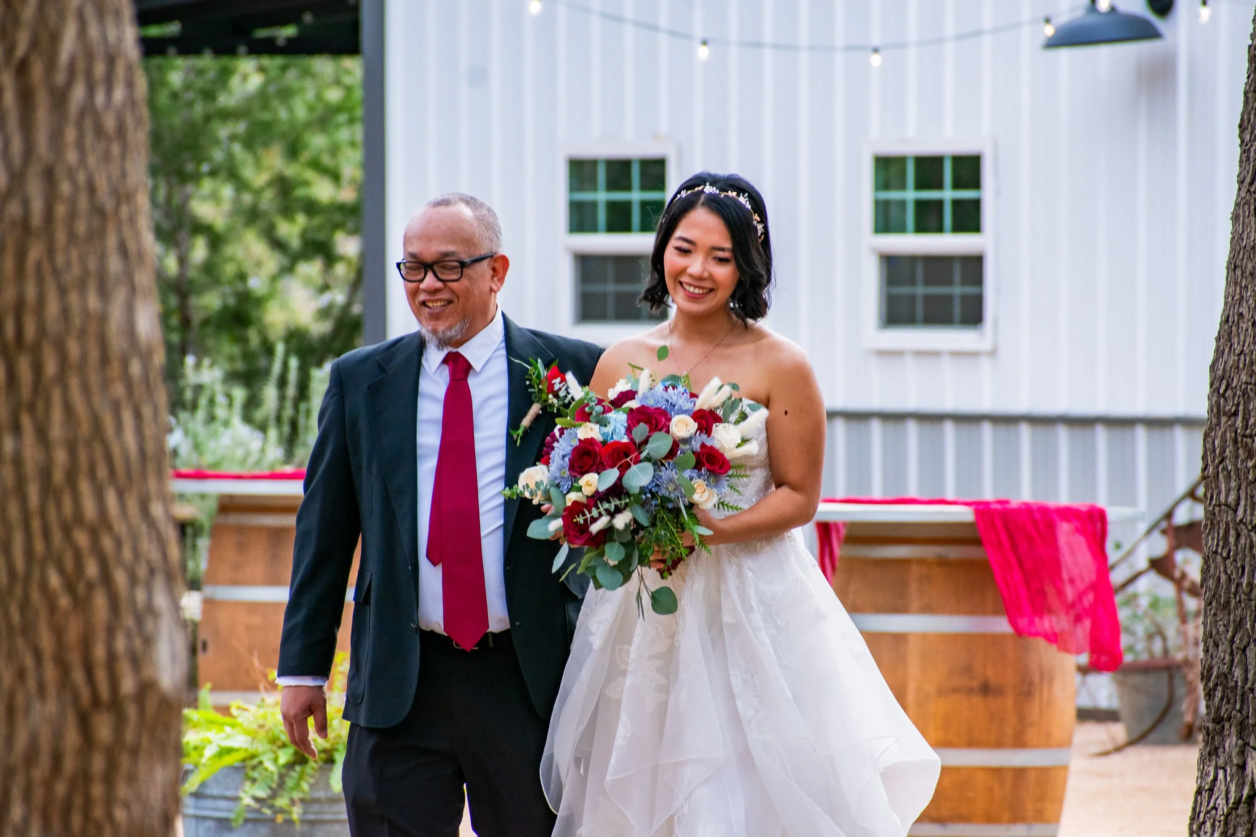A bride and her father walking outdoors during a wedding ceremony, with the bride holding a colorful bouquet of flowers and smiling.