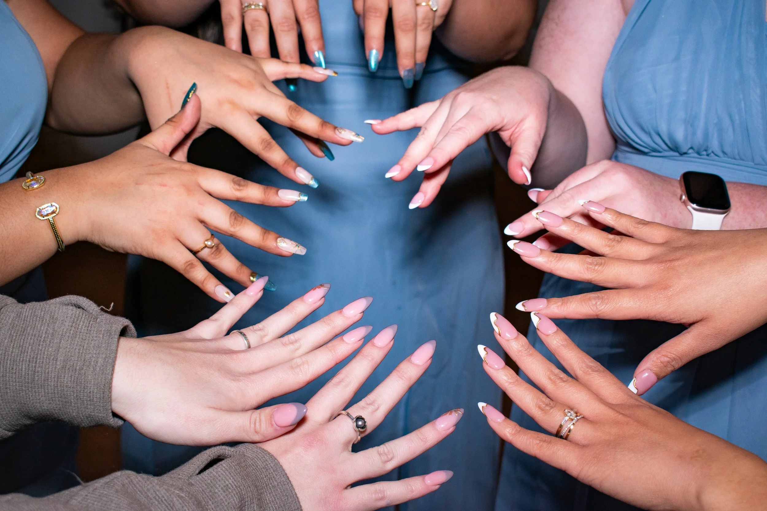 Multiple hands reaching towards the center, displaying various manicured nails, jewelry, and a smartwatch. The nails are decorated with different designs and colors.