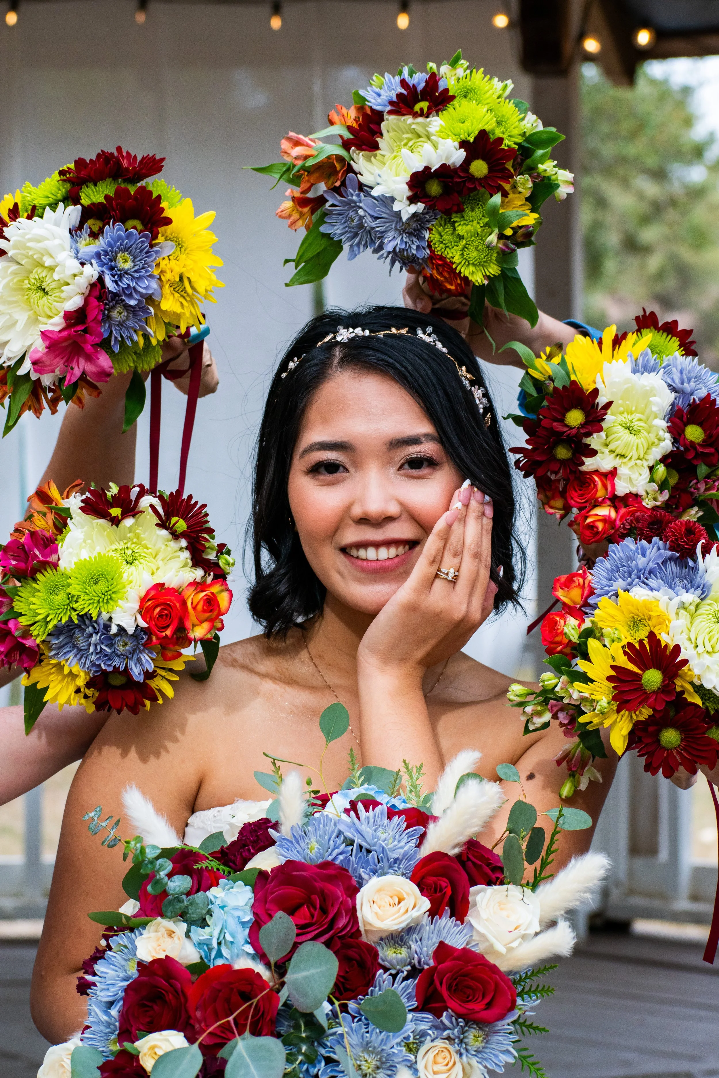 Young woman with dark hair wearing a floral headband and a strapless dress, holding a bouquet of flowers and surrounded by additional floral arrangements, outdoors with a wooden structure in the background.
