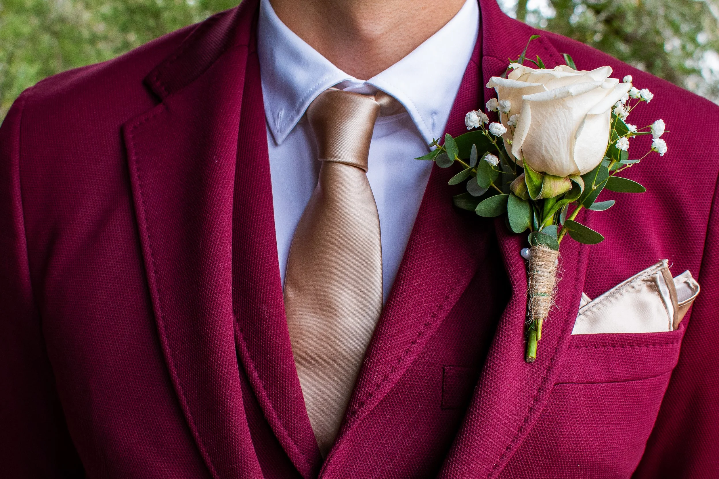 Close-up of a man wearing a burgundy suit jacket, white dress shirt, and a beige tie, with a boutonniere of white roses and small white flowers pinned to the jacket's lapel.