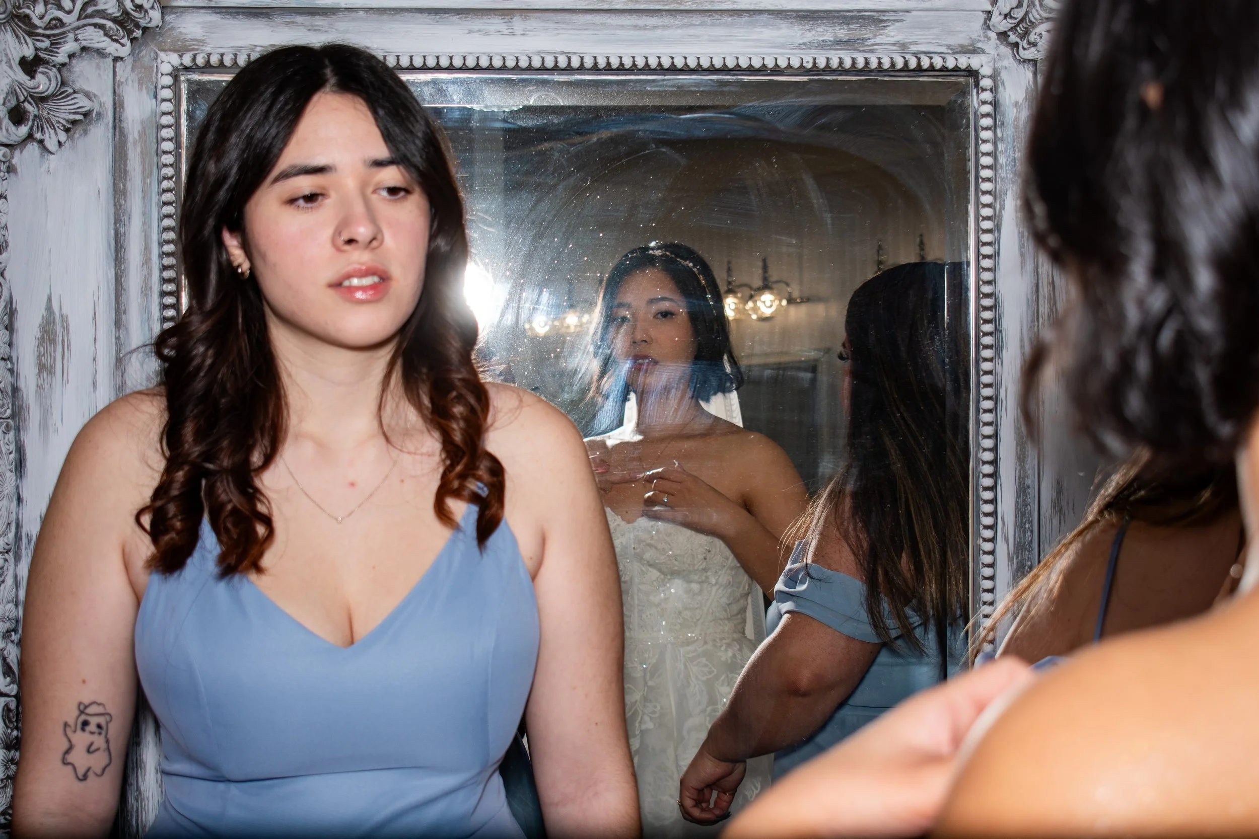 A woman with dark hair and a blue dress looking at her reflection in a mirror, with her reflection visible in the mirror.