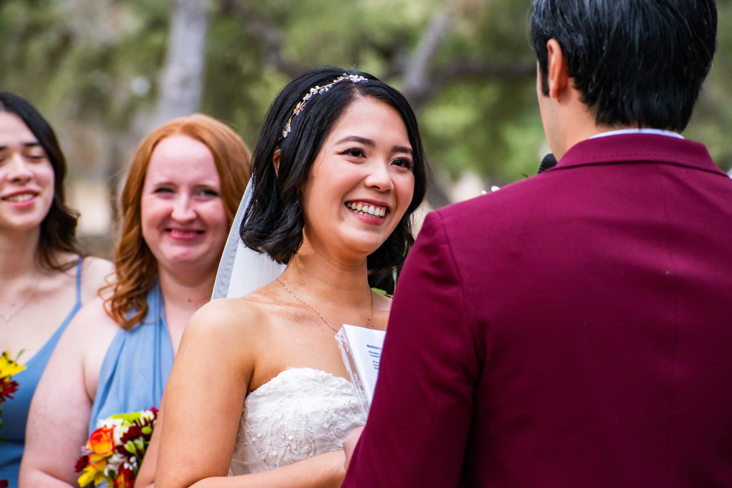A bride in a white wedding dress smiling at her groom during their wedding ceremony outdoors, with two bridesmaids visible in the background holding bouquets.
