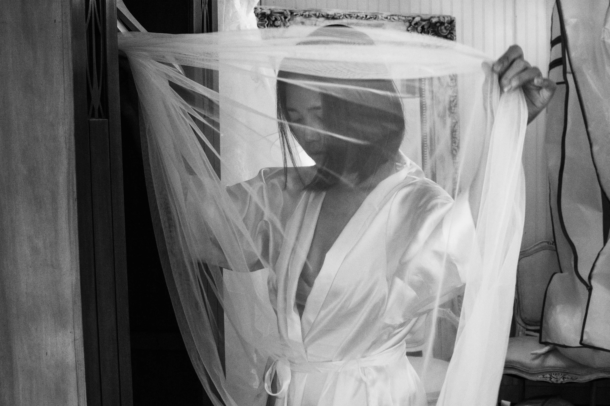 A woman wearing a white garment and a veil, holding the veil with her left hand, with her face partially seen through the veil. She's indoors with a wall, curtain, and a chair nearby.