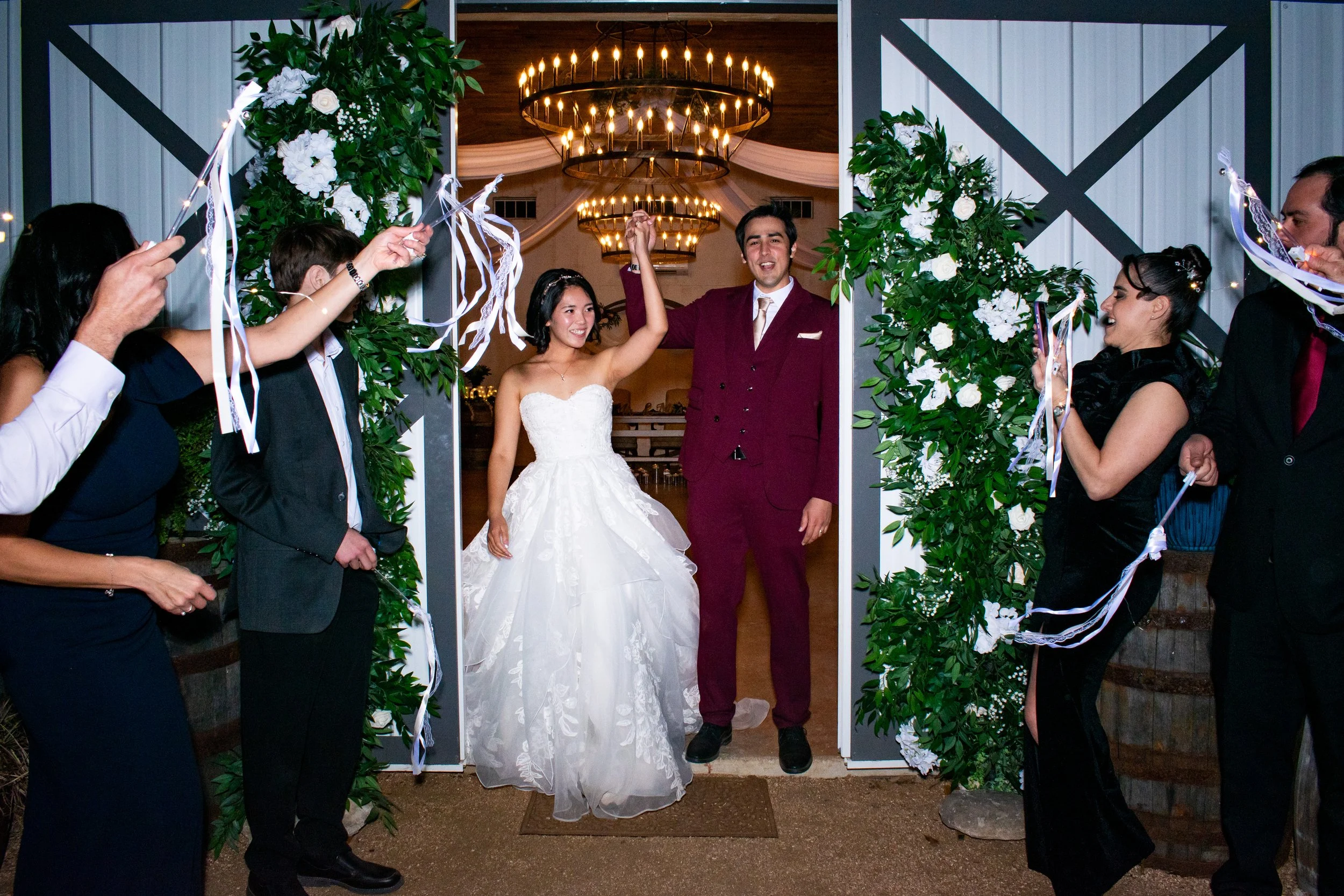 A wedding celebration scene with a bride and groom standing under a decorated archway, surrounded by friends holding sparklers, inside a venue with chandeliers and draped curtains.