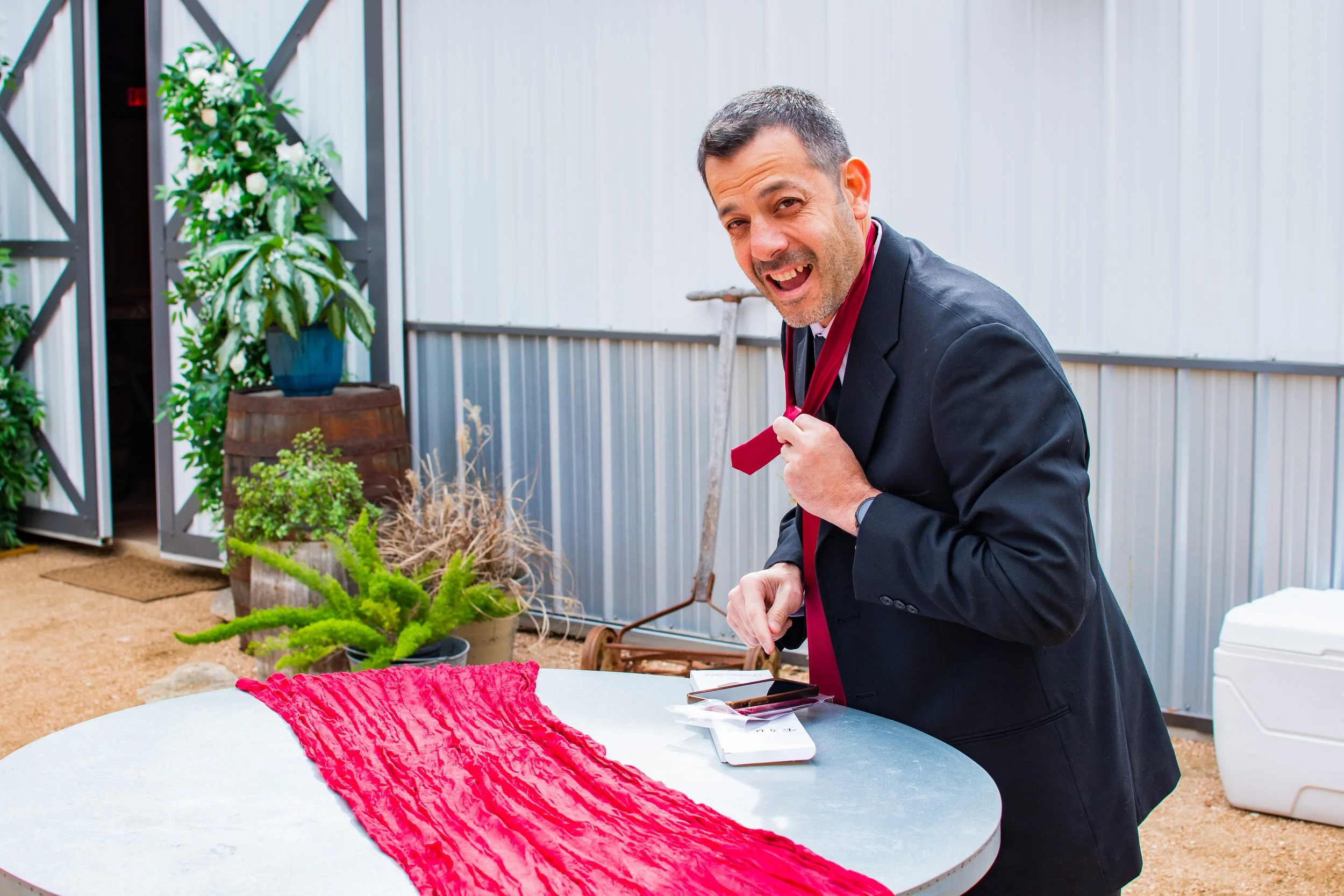 A man in a suit with a red ribbon around his neck stands at a table, smiling and adjusting the ribbon. The table has a red fabric centerpiece, and balloons, flowers, and plants are visible in the background.