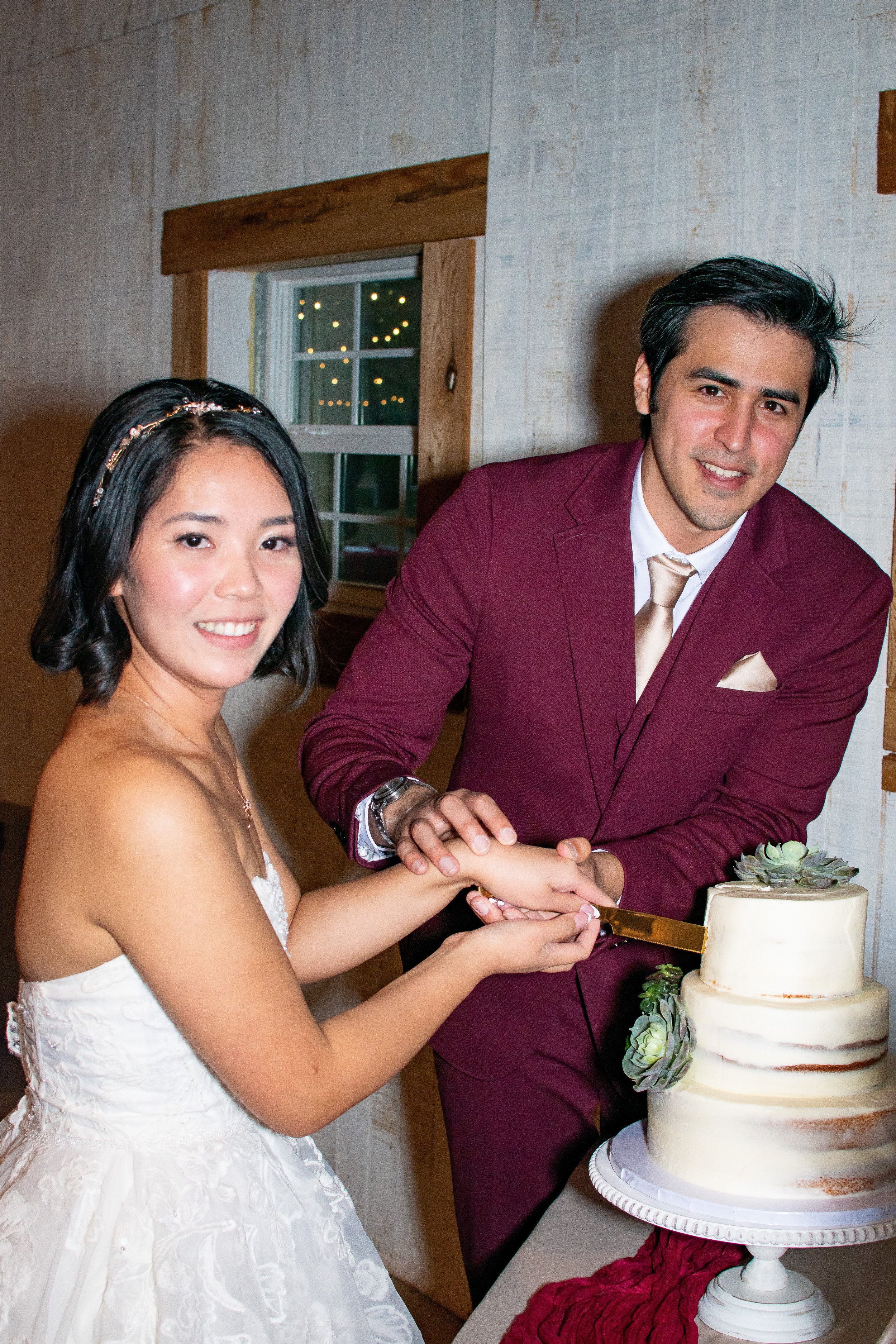A bride and groom cut a wedding cake together. The bride wears a white lace wedding dress, and the groom wears a maroon suit with a white shirt and cream-colored tie. They are smiling and holding the cake knife, which is gold. The wedding cake is a t