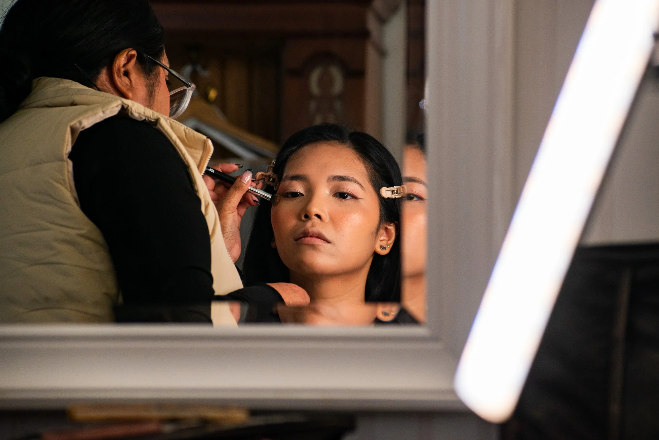 Makeup artist applying makeup to a young woman with closed eyes, sitting in a mirror, with a focus on her face.