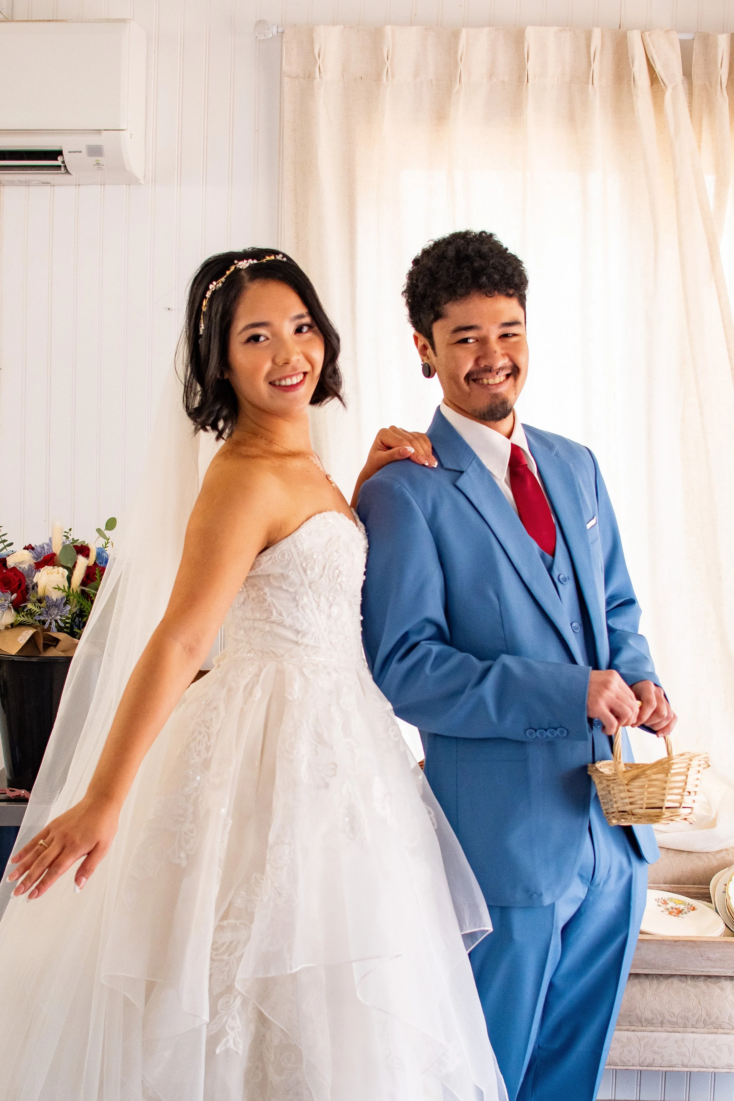 A bride and groom smiling, standing side by side indoors in front of beige curtains, with the bride wearing a strapless white wedding gown and the groom in a blue suit holding a basket.