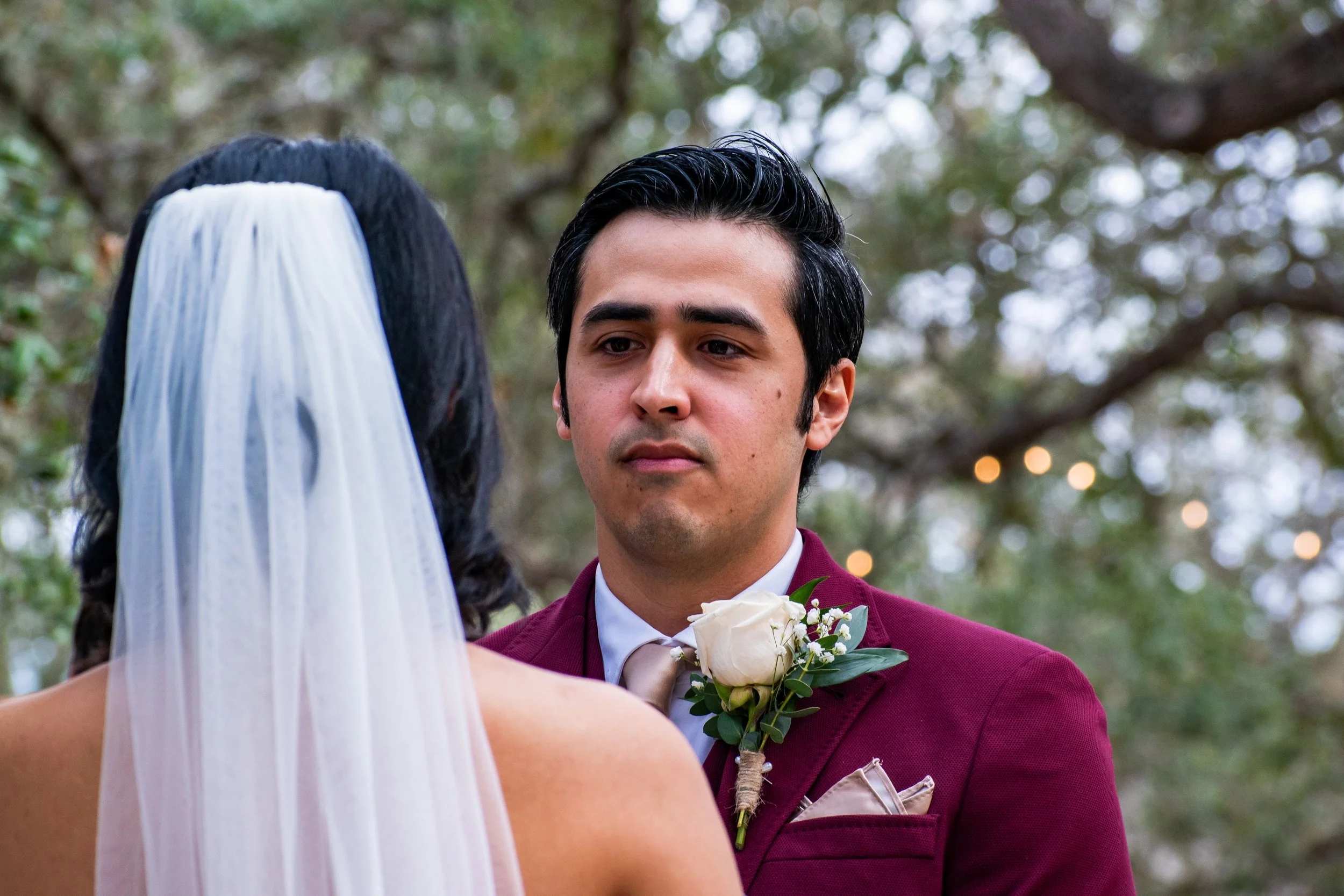 A man in a maroon suit and white shirt weds a woman with a white veil during an outdoor wedding ceremony.
