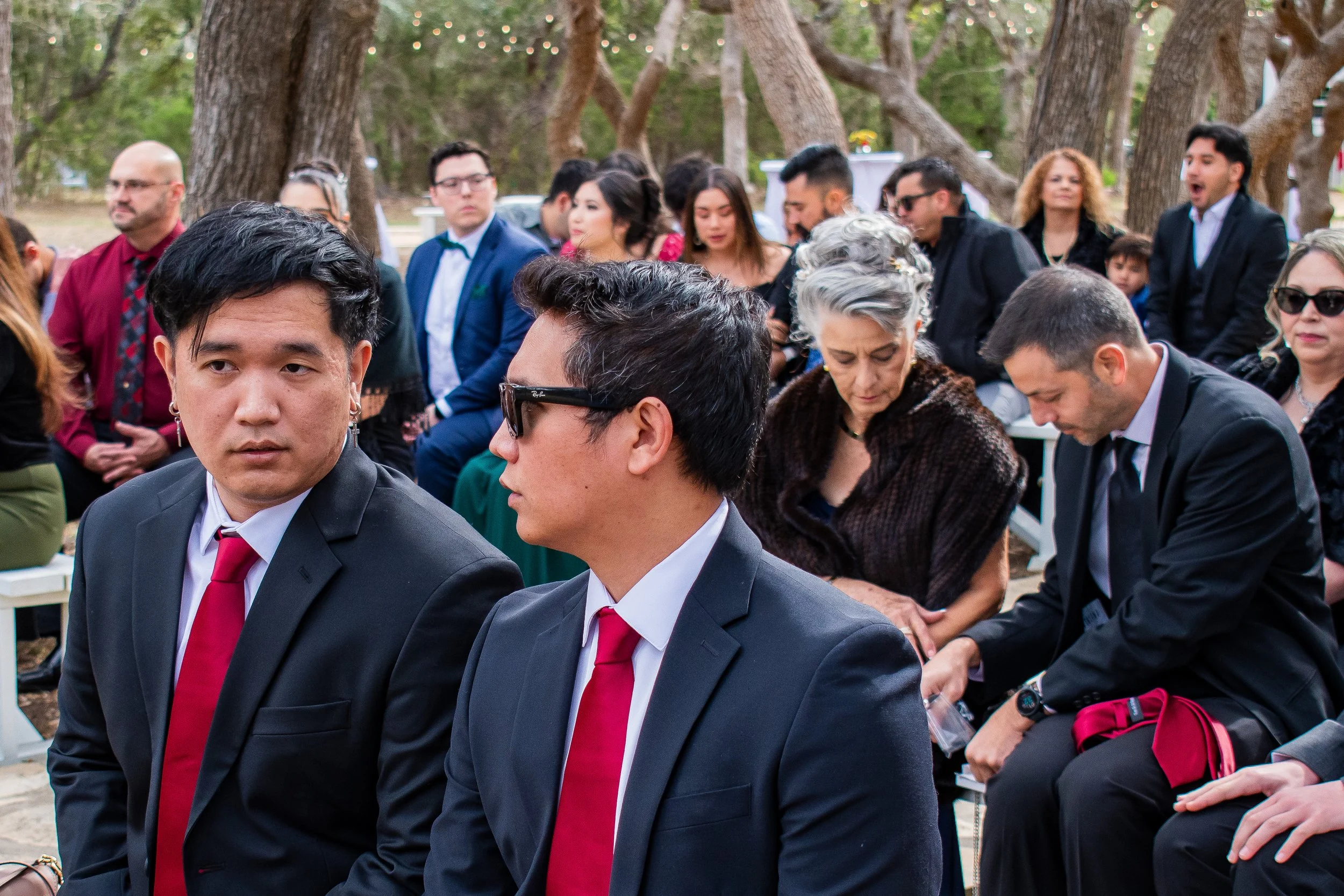 A group of diverse people dressed in formal attire gathered outdoors, sitting closely together with trees in the background, possibly at a wedding or formal event.
