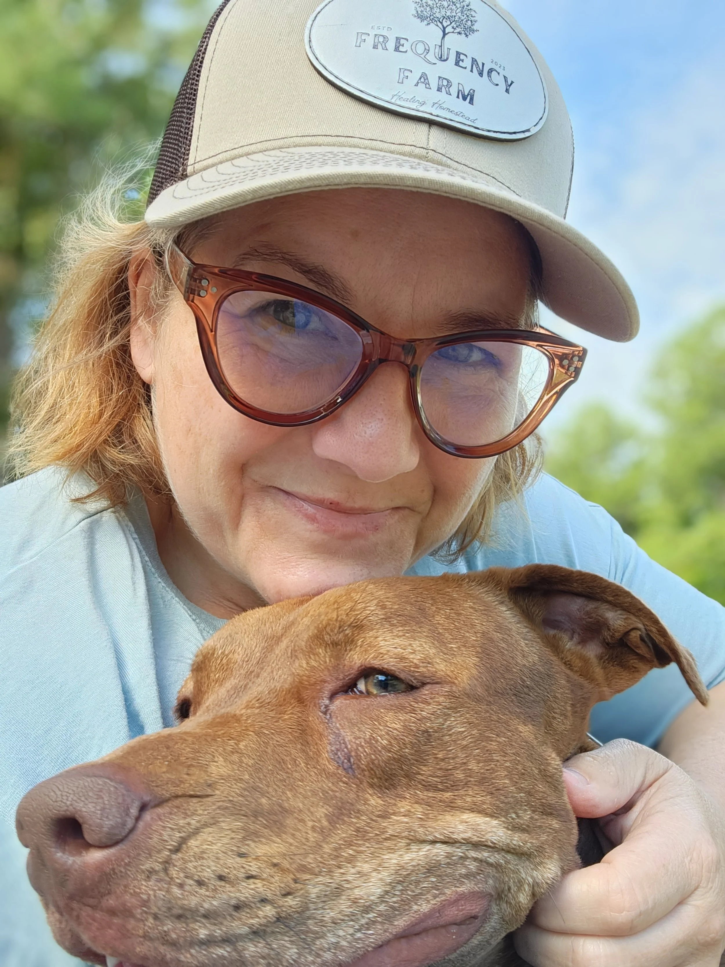 Woman wearing sunglasses and a beige cap with a 'Frequency Farm' patch, hugging a brown dog outdoors. The background has trees and a blue sky.