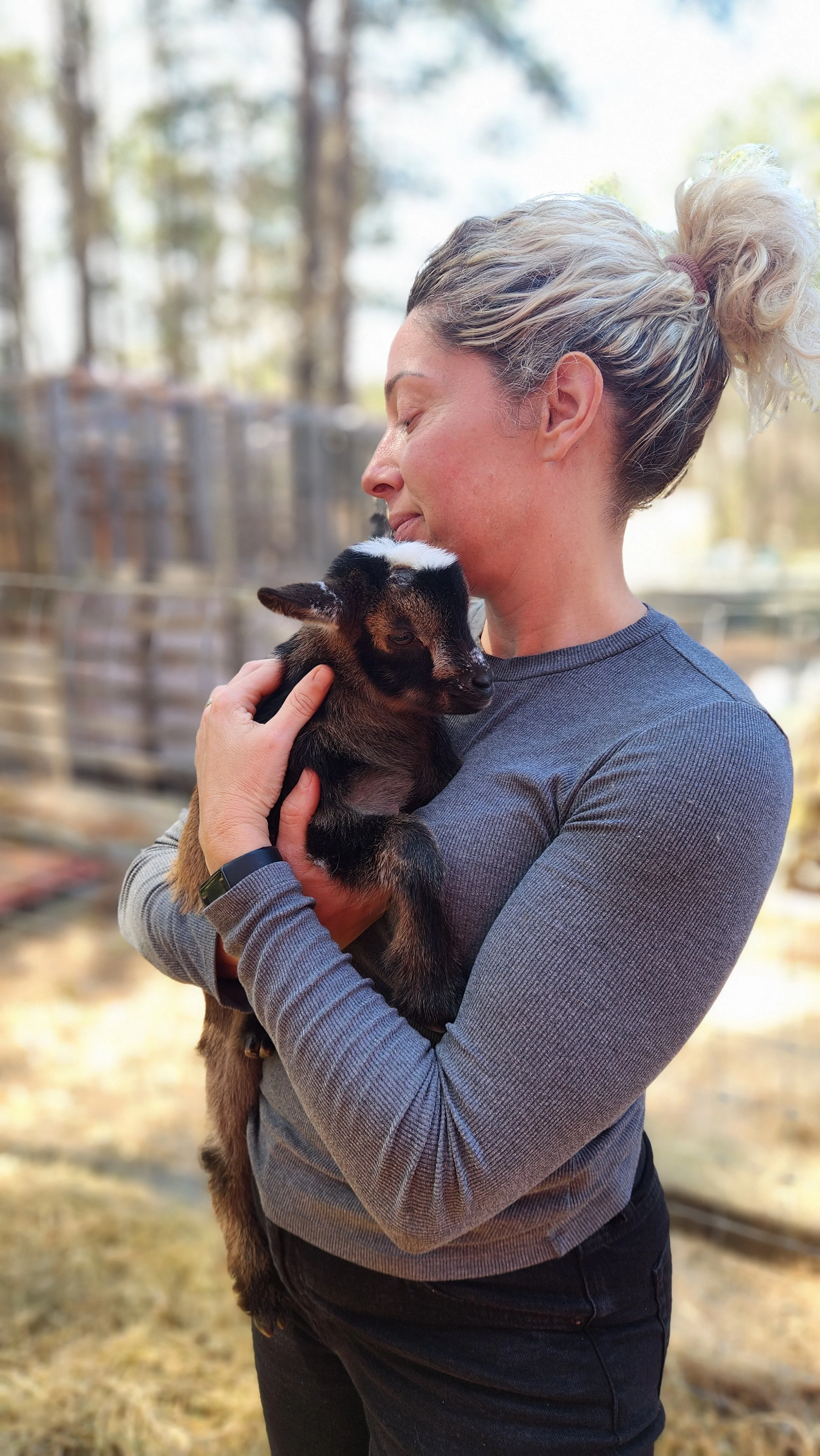 A woman holding a baby goat outdoors, with trees and a wooden fence in the background.
