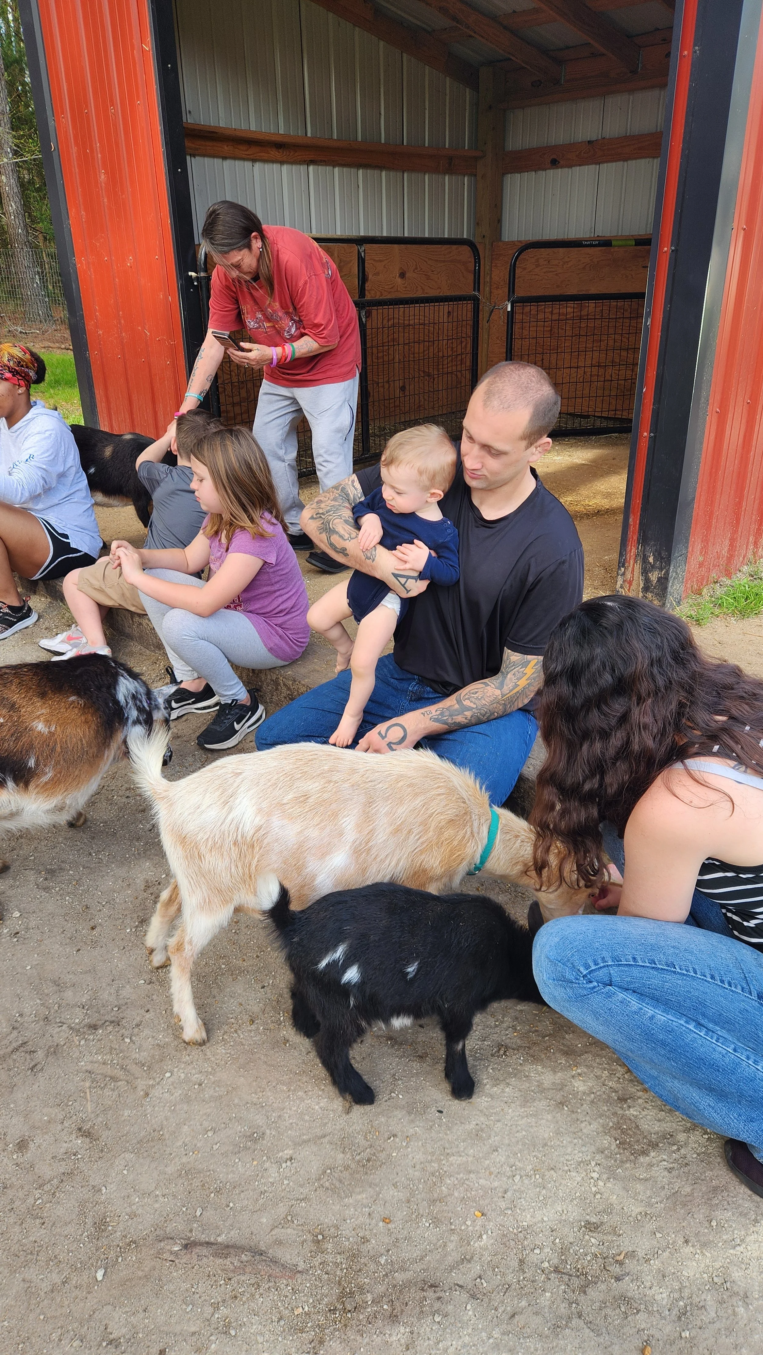 Group of people, including children and adults, interacting with goats inside a barn or petting zoo area. A man with tattoos holds a young child, and a woman is feeding a black goat while children sit nearby.
