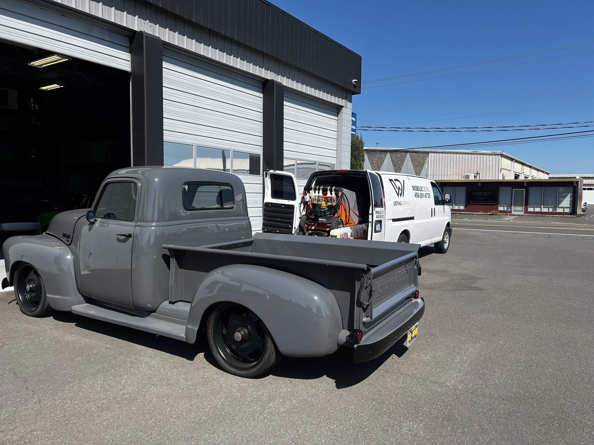 A vintage gray pickup truck parked outside a building with its bed filled with equipment. A white service van with the company's logo and contact information is parked nearby.