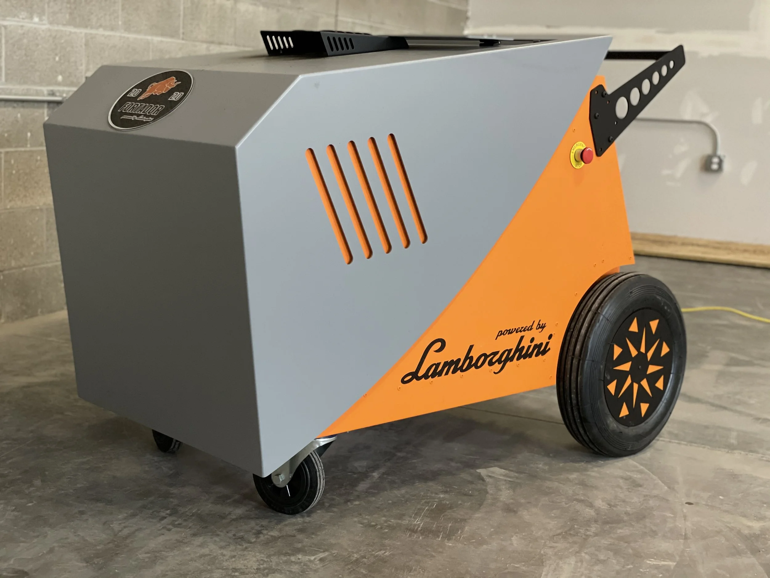 A large, gray, orange, and black industrial machine with wheels, labeled "powered by Lamborghini," placed on a concrete floor in front of a cinder block wall.