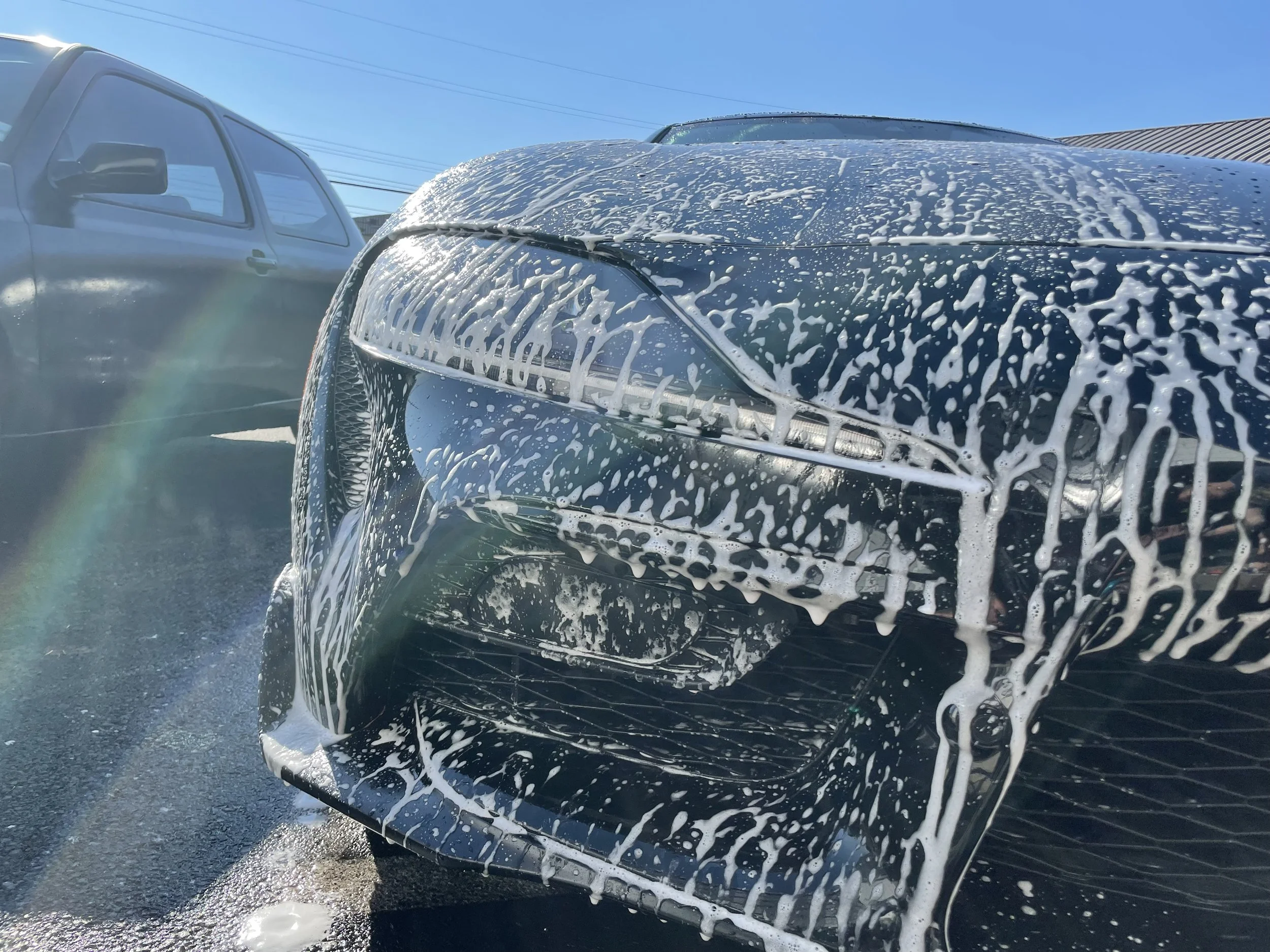 Close-up of a black car being washed with soap, with foam on the front, and another vehicle visible in the background on a bright, clear day.