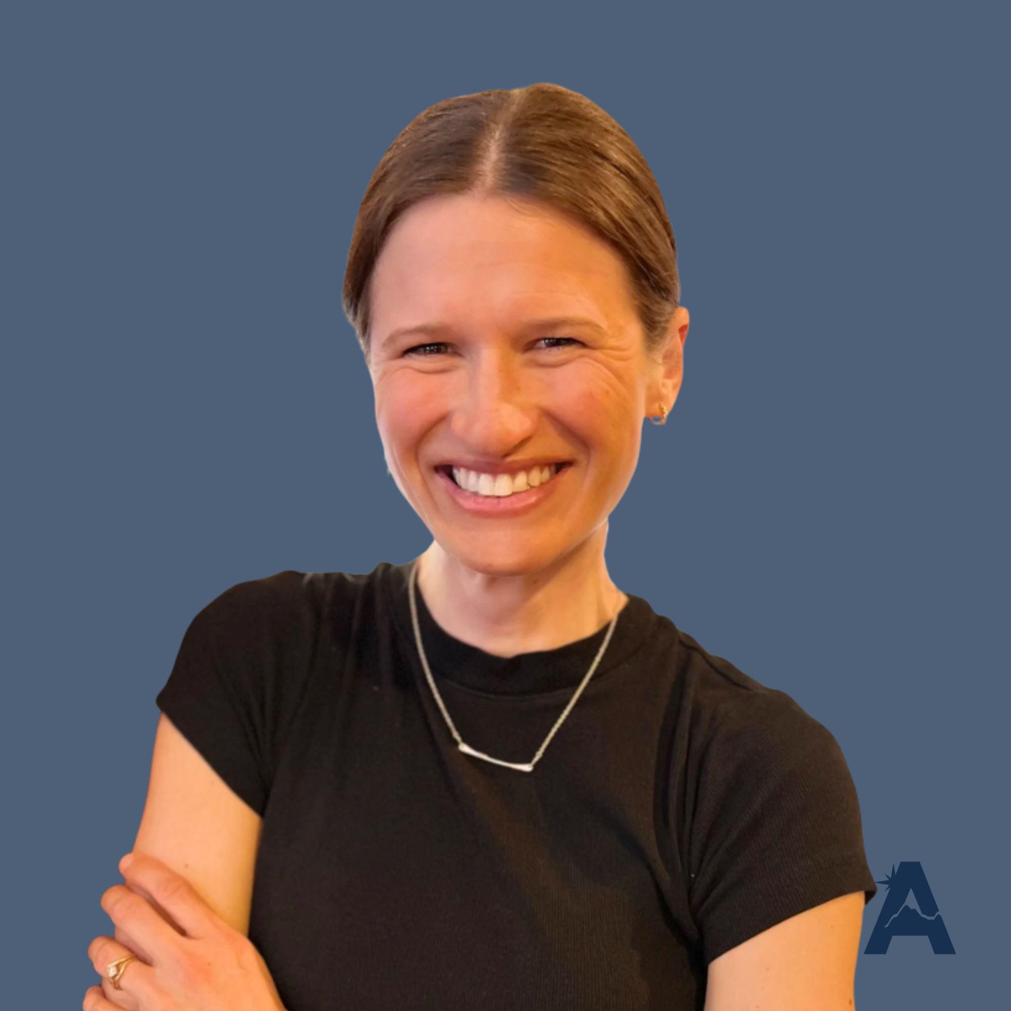 A woman with brown hair smiling and leaning on a wooden post, wearing a black t-shirt and jewelry, in a warmly lit indoor setting.