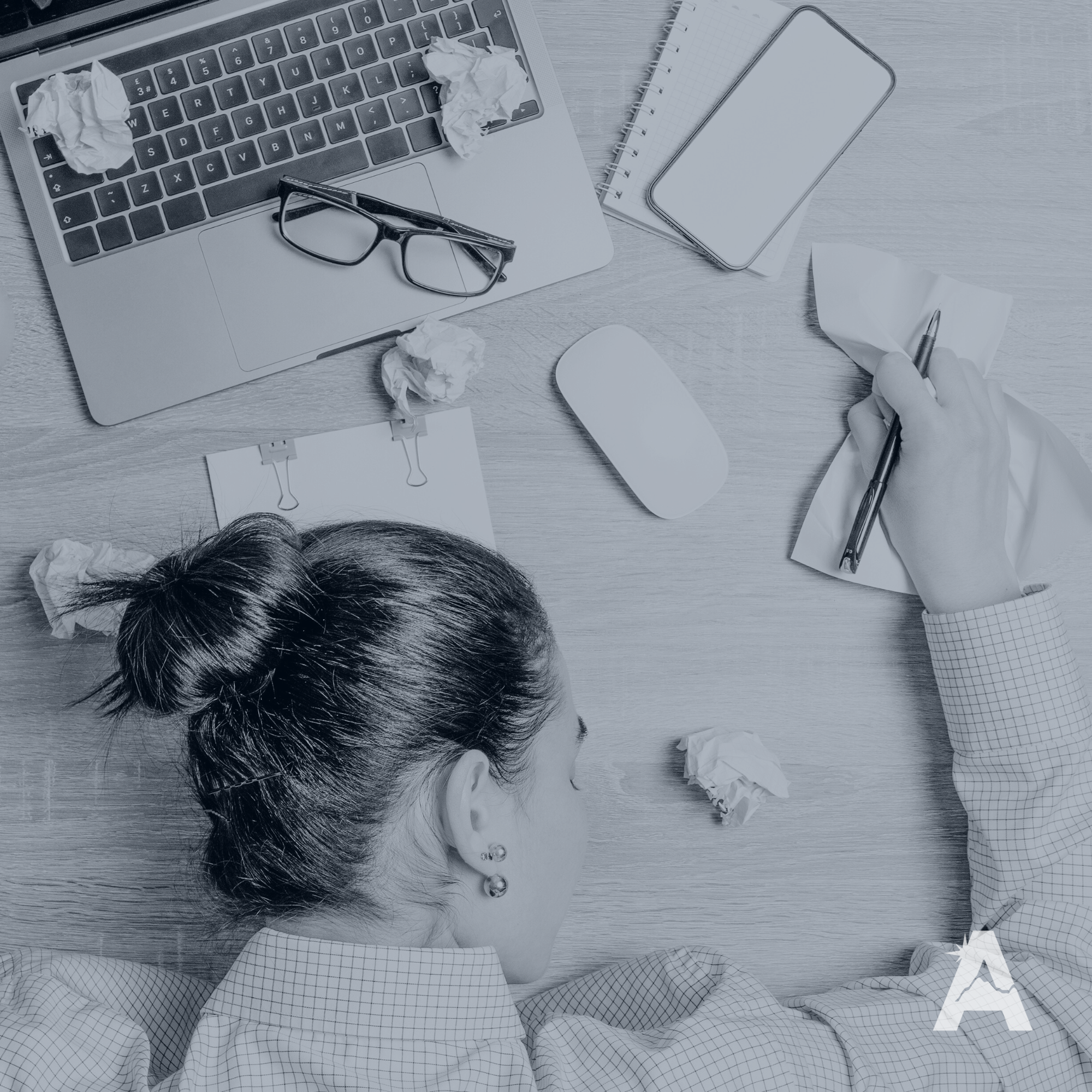 Overhead view of a woman at a cluttered desk with crumpled paper, a laptop with glasses on top, a wireless mouse, a notebook, a smartphone, and a person writing on a notepad.