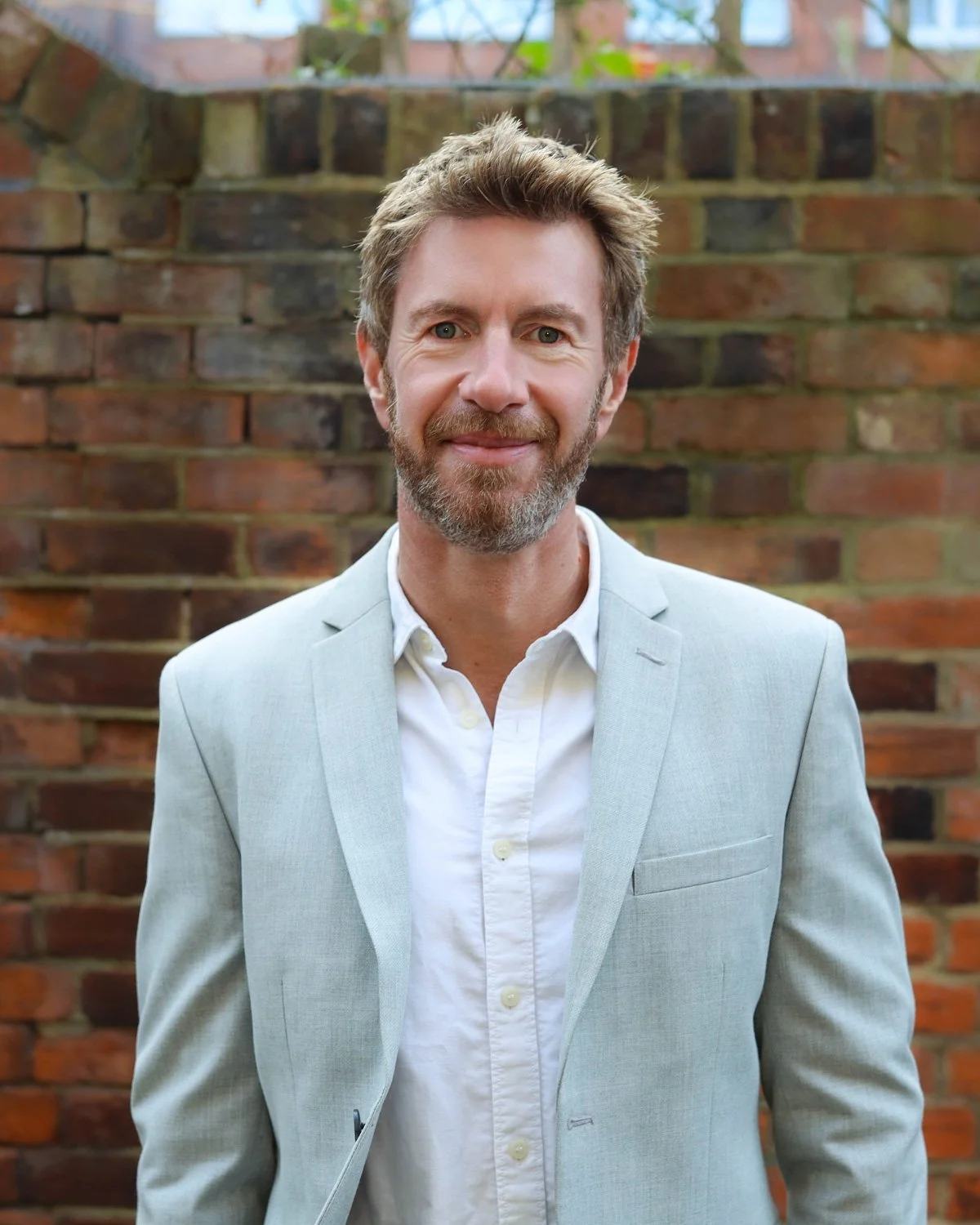 A man with light brown hair and a beard wearing a dark gray polo shirt against a grayish background.