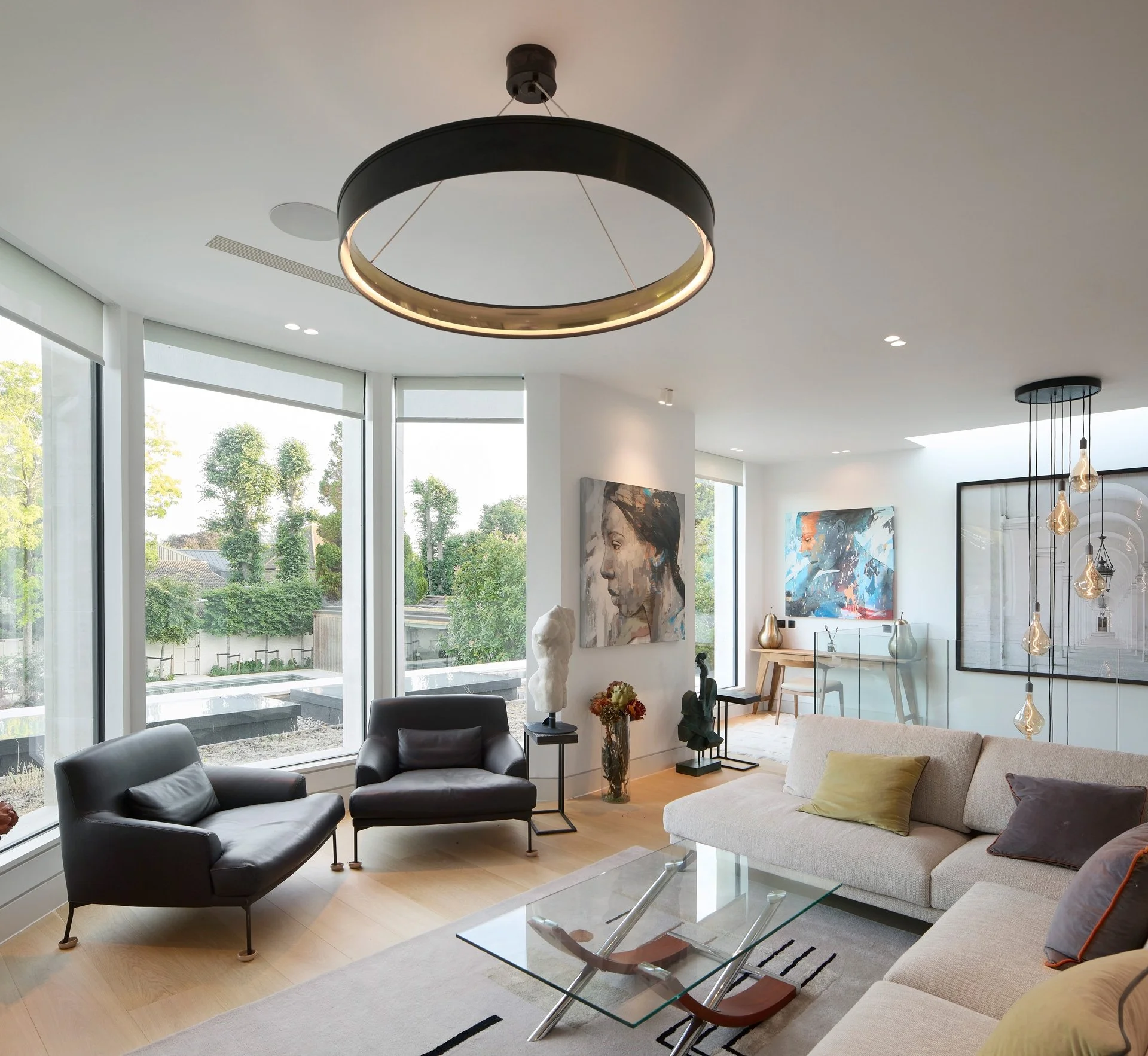 Modern living room with large windows, black and beige sofas, glass coffee table, contemporary art, and unique hanging light fixtures.