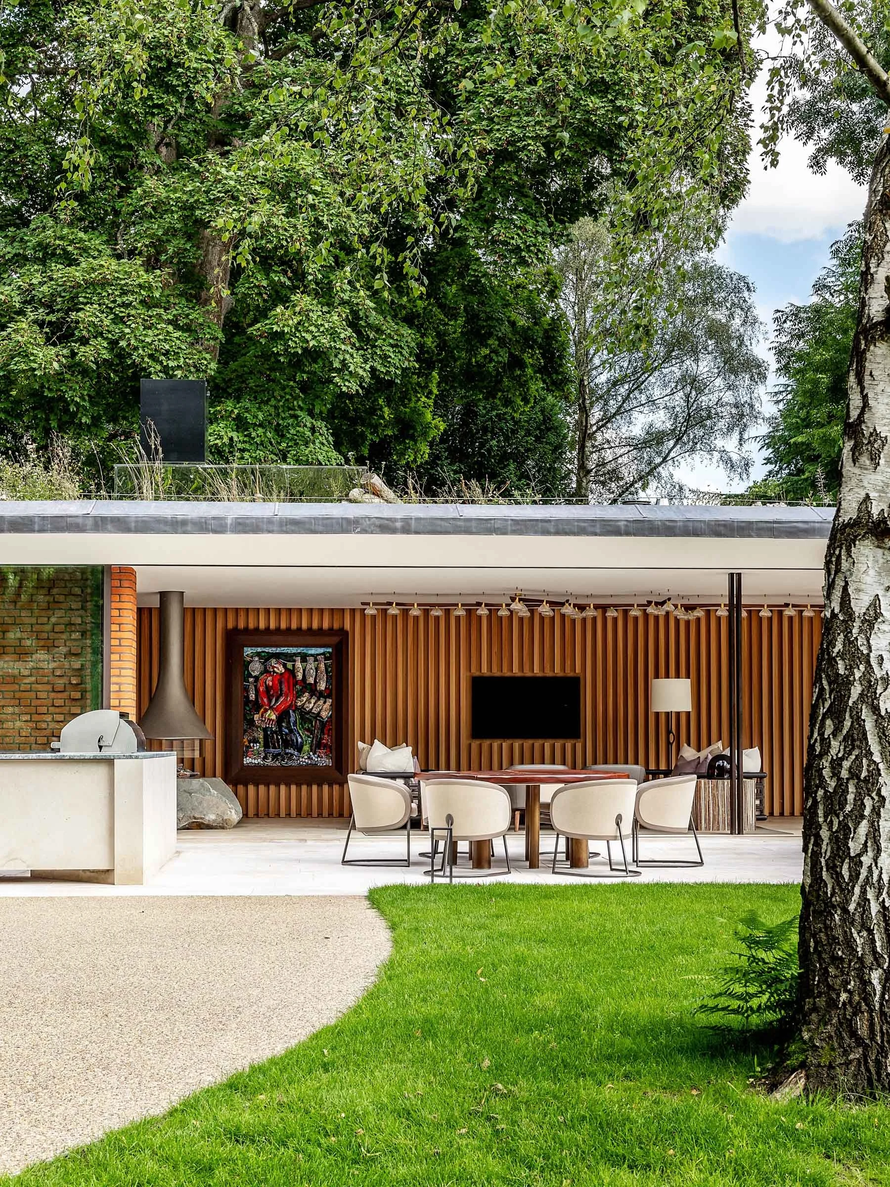 Modern outdoor patio with a round dining table and chairs, a flat-screen TV, wooden slat wall, and lush green trees in the background.