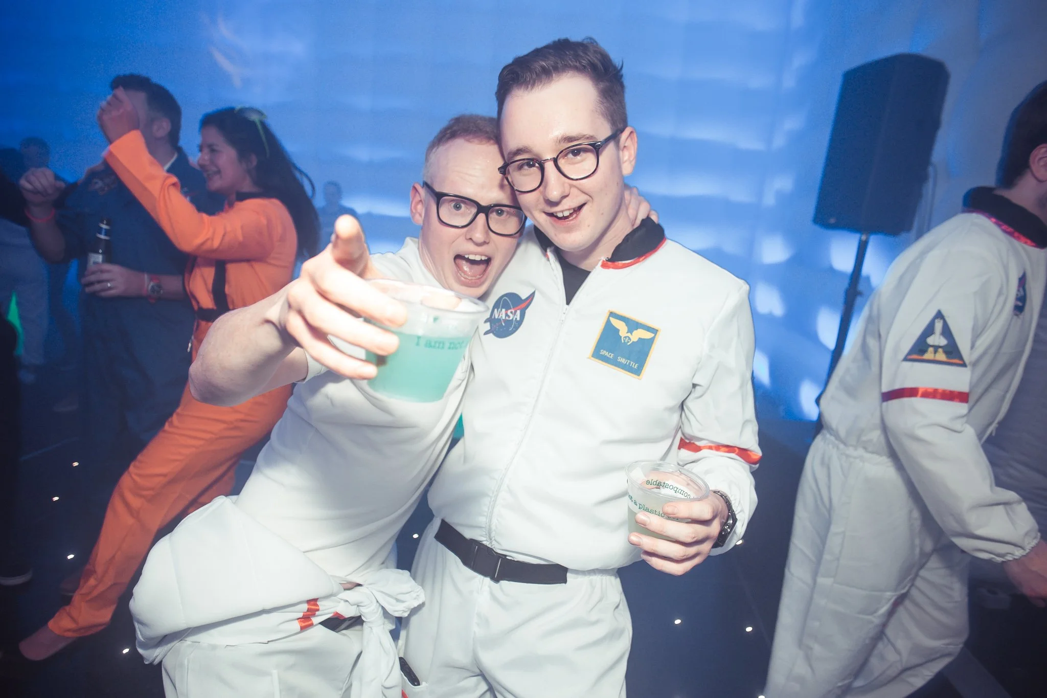 Two men dressed as astronauts celebrating at a party, one holding a drink and the other with his arm around his friend, with other party attendees in the background.