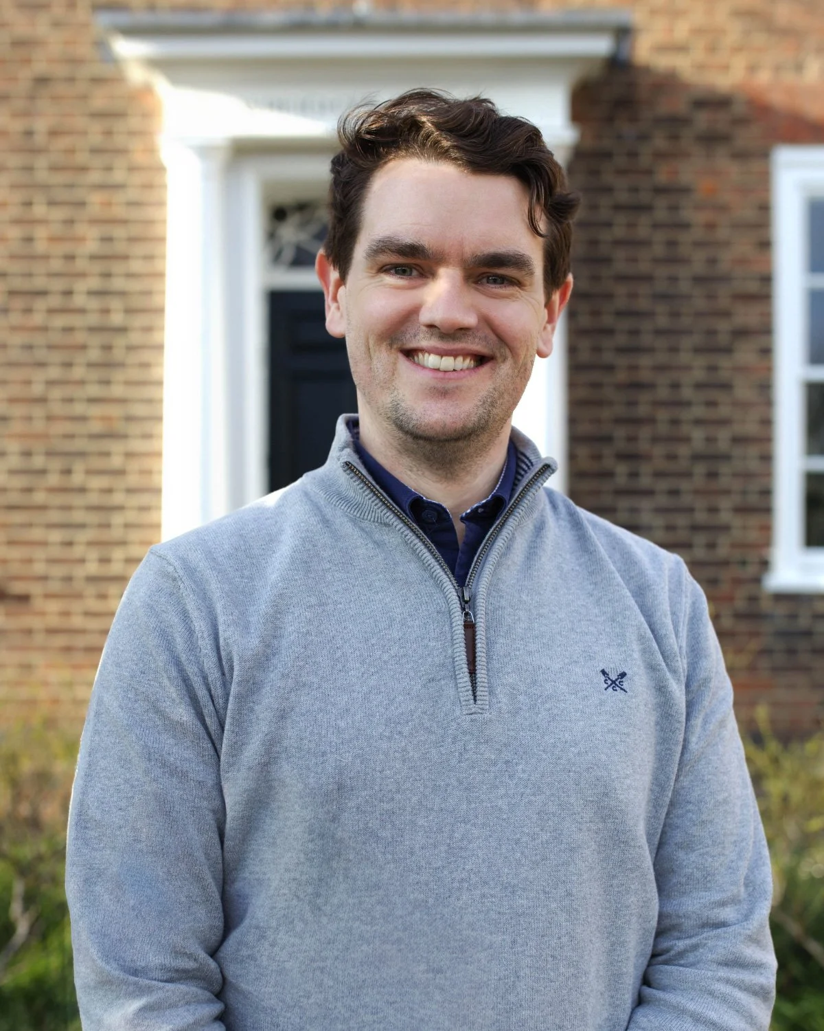 Portrait of a man with dark hair, wearing a light-colored button-up shirt, against a plain blue background.