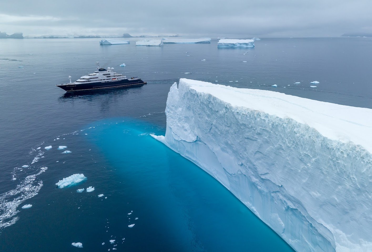 A large iceberg with a flat top floating in the ocean, with a luxury yacht nearby and more icebergs visible in the background.