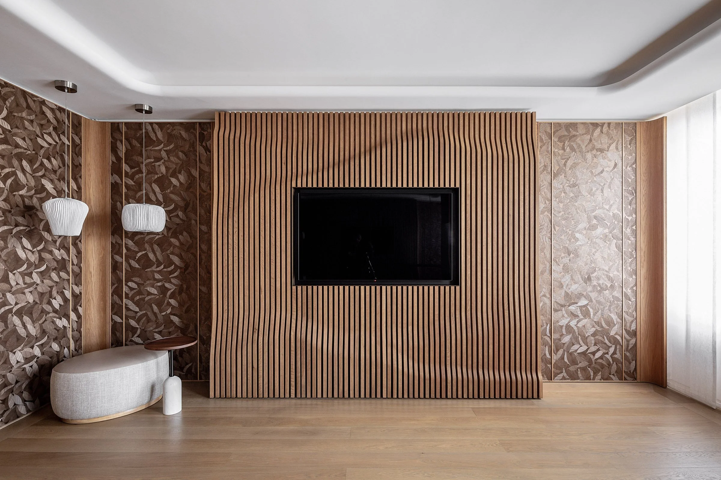 Modern living room with a black flat-screen TV mounted on a vertical wooden slat wall, surrounded by patterned wallpaper on both sides, with two white pendant lights, a small beige cushioned bench, and light wooden flooring.
