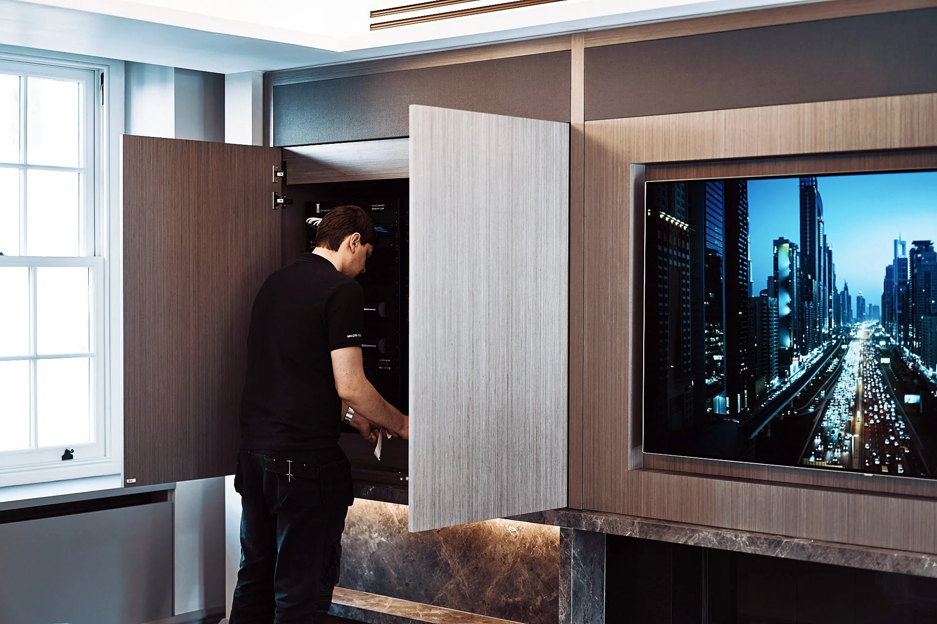 A man in black clothing opening a server rack cabinet in a modern office or conference room, with a large cityscape image on the wall.