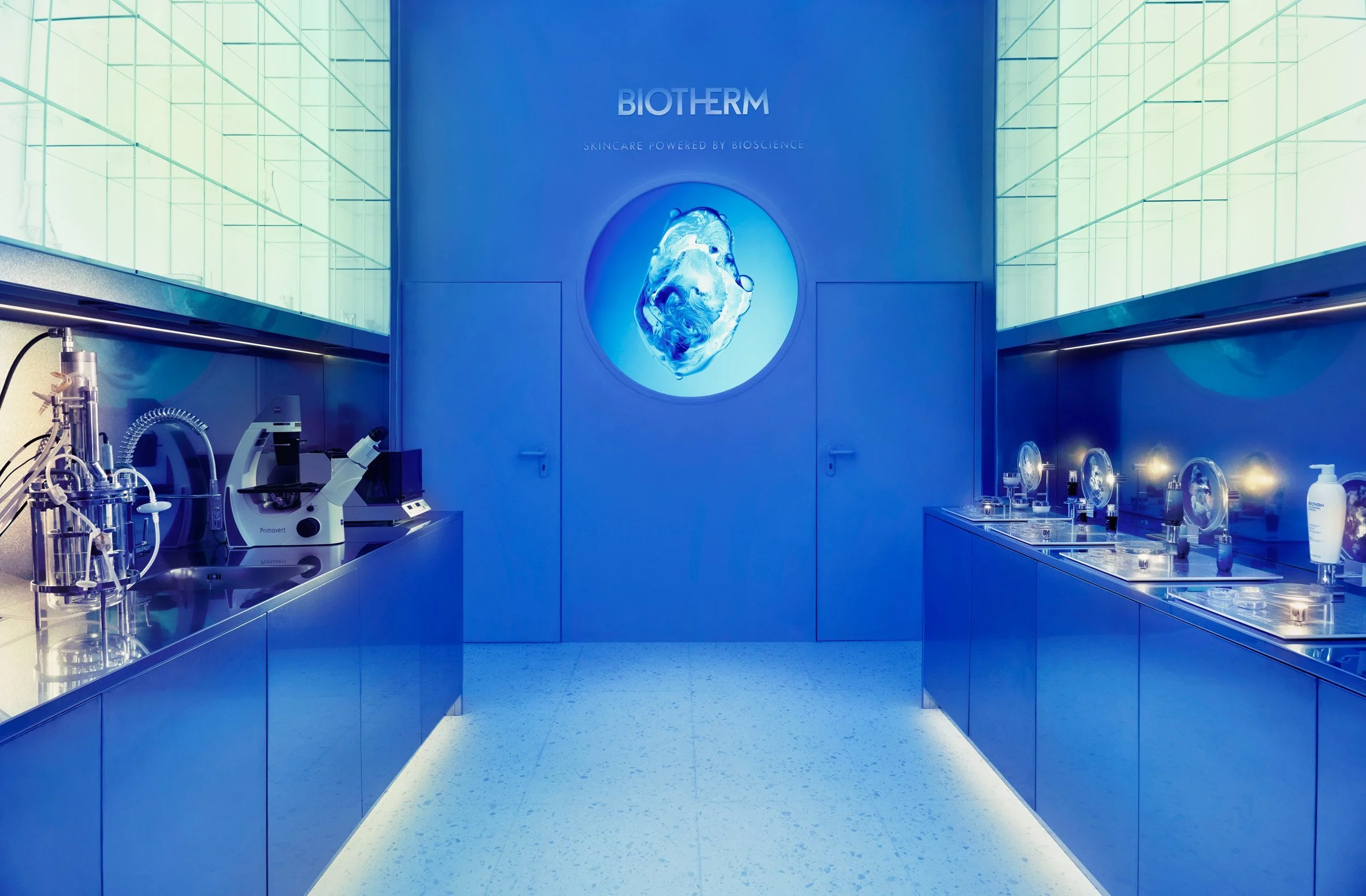 Interior of a modern skincare lab with blue lighting, featuring microscopes and skincare products, and a Biotherm logo on the wall.