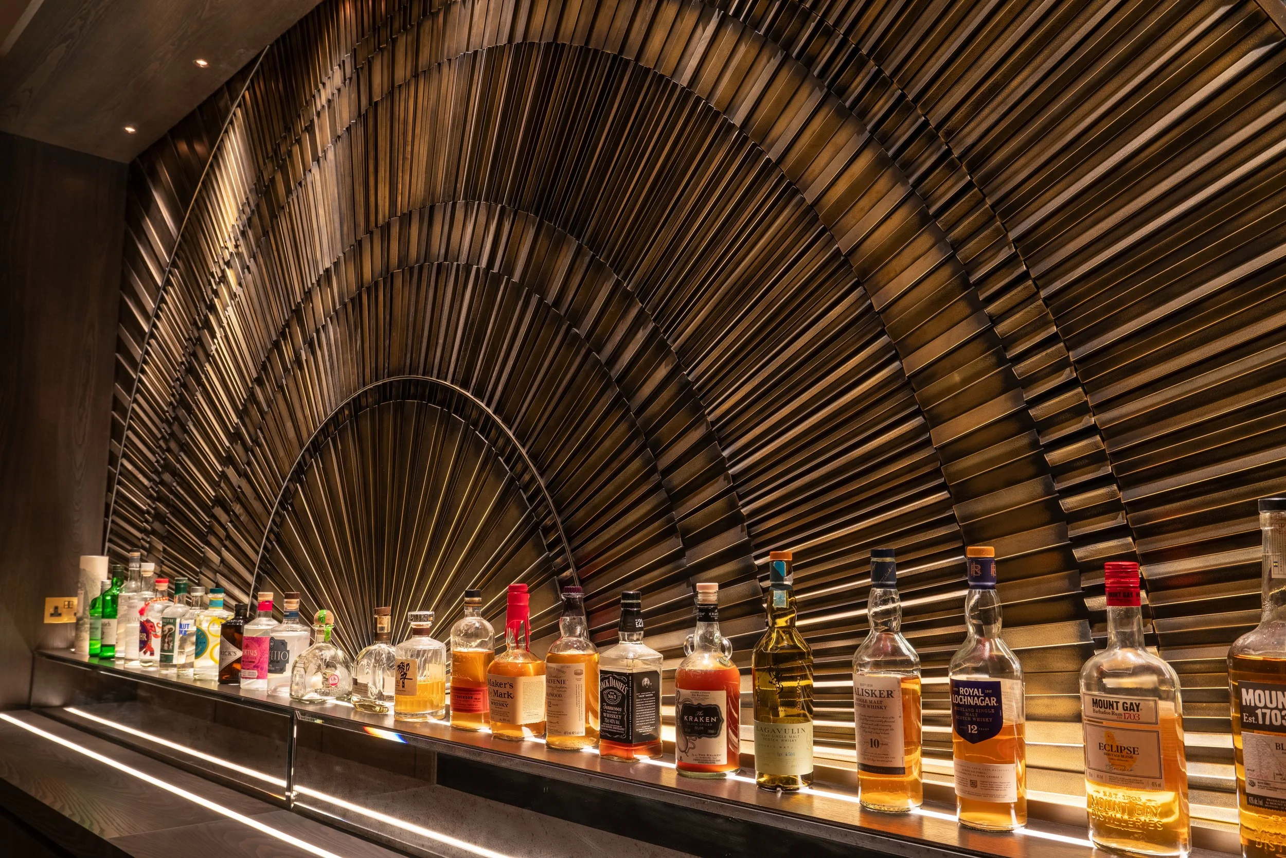 A selection of liquor bottles arranged on a shelf with a decorative, radiating metal wall design in the background.