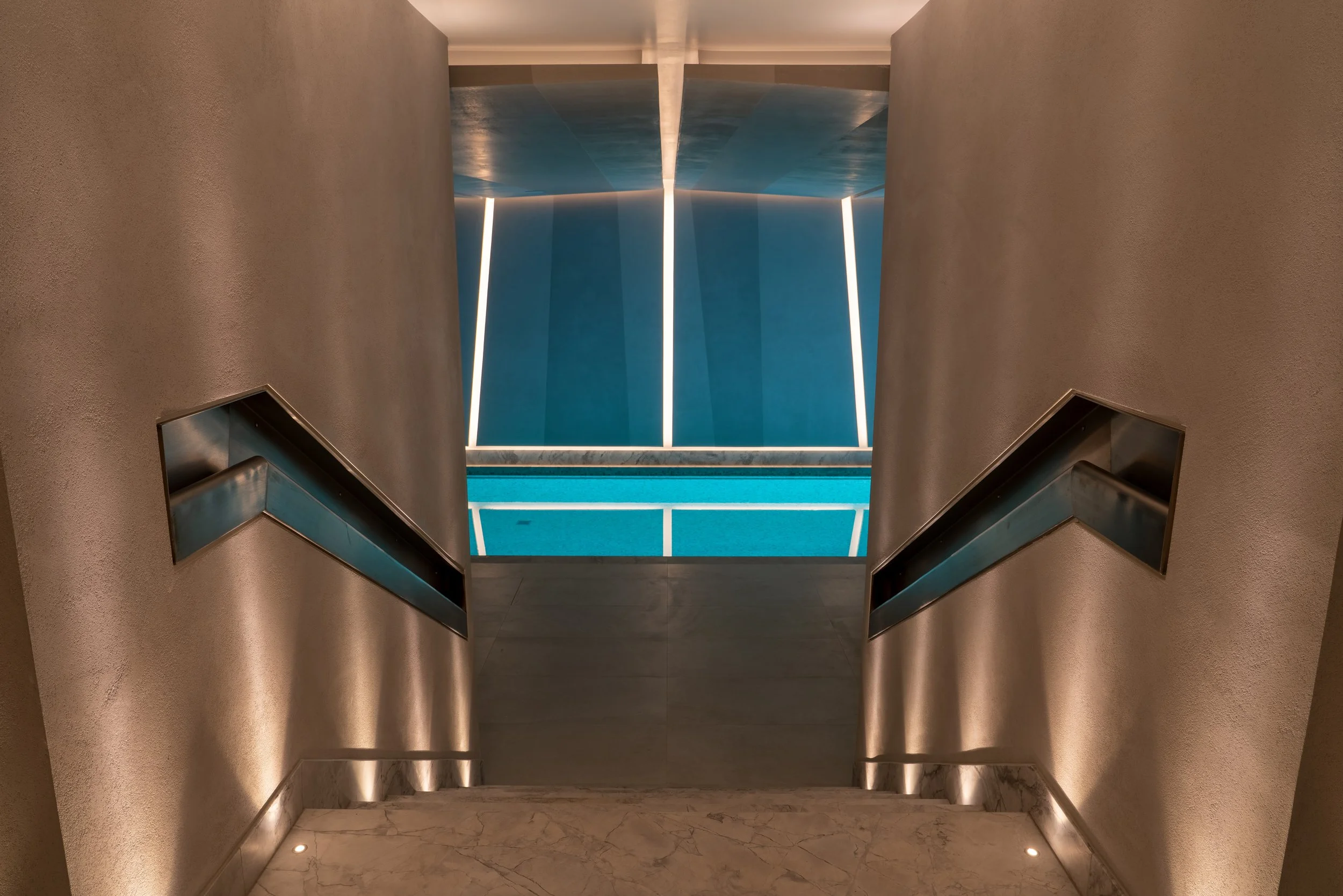 Indoor staircase leading down to a swimming pool with blue water, illuminated by modern vertical light fixtures and recessed lighting, with marble flooring at the top of the stairs.