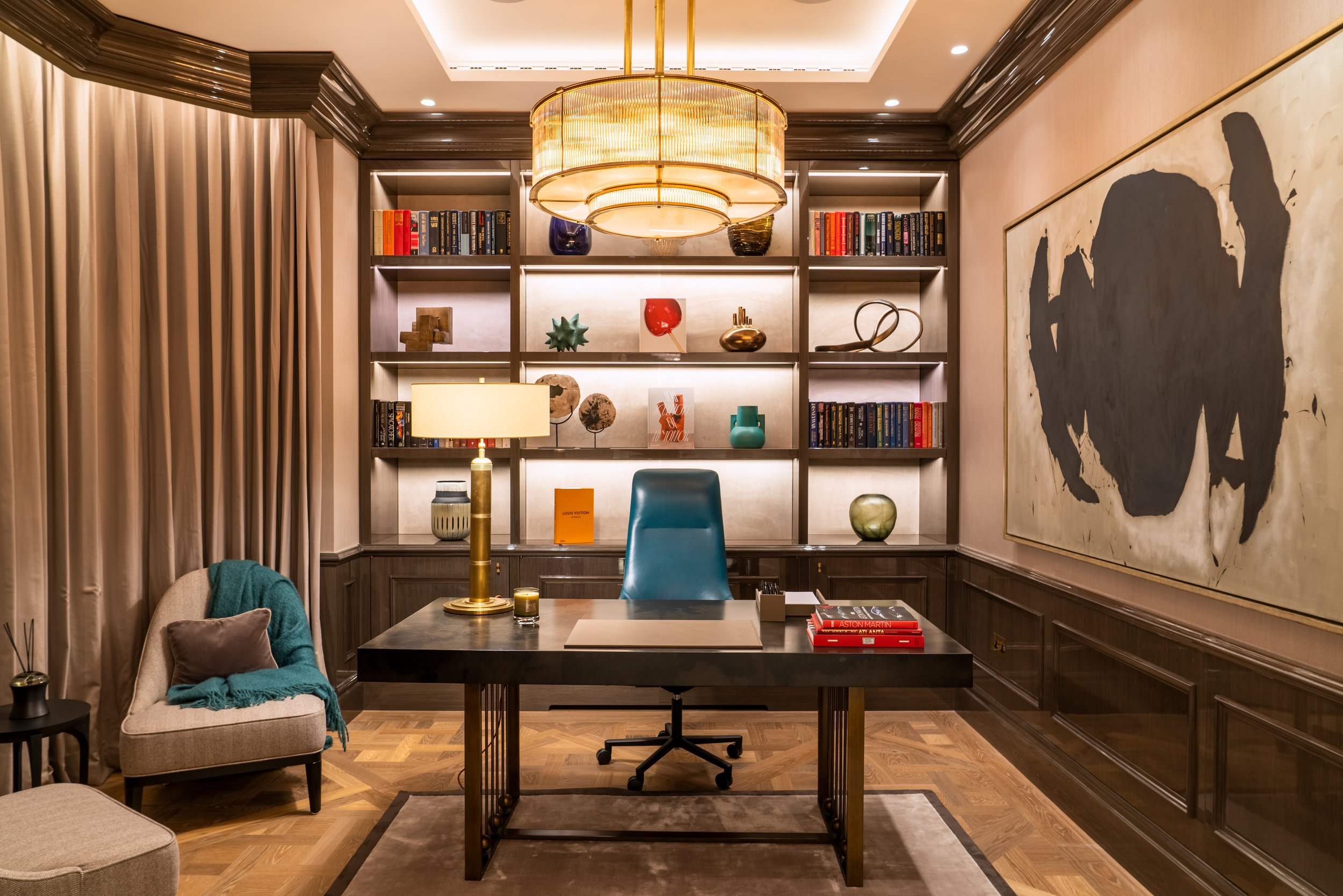 A luxurious home office with a dark wood desk, green office chair, beige armchair with a pillow and blue throw, large abstract painting on the wall, built-in bookshelves with colorful decorative items, a modern chandelier, and warm lighting.