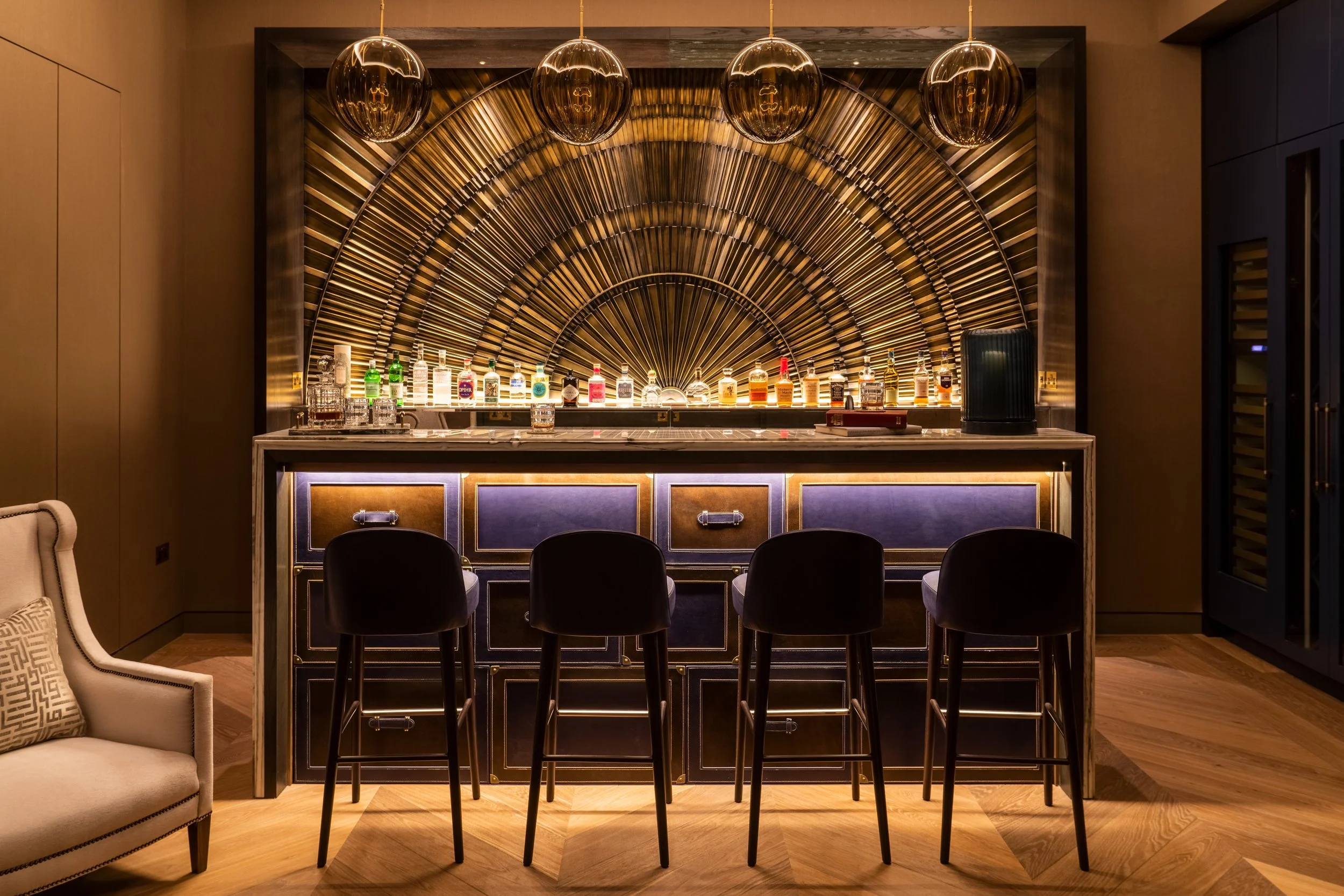 A luxurious bar with a dark purple and gold decorated counter, four black bar stools, a beige armchair with a patterned pillow to the left, and a backlit golden decorative wall behind the bar with hanging gold lights.