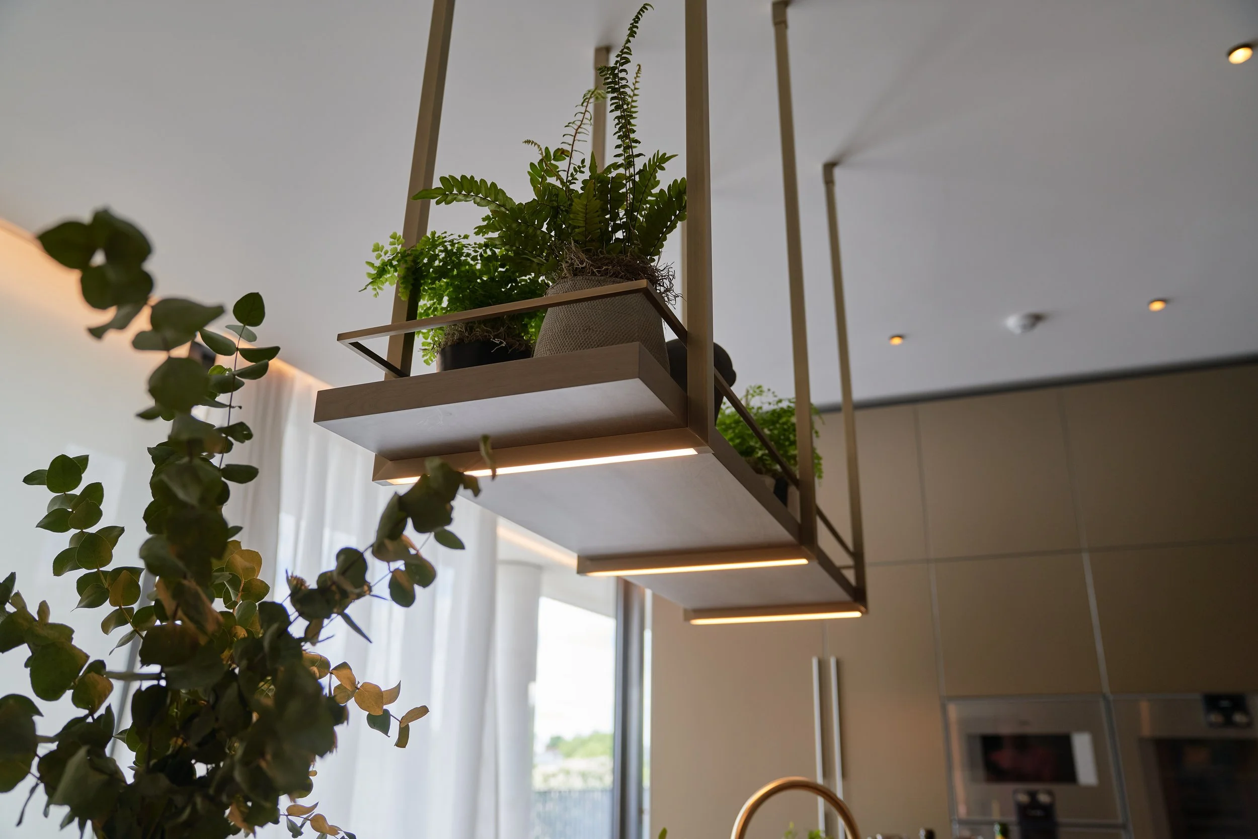 Indoor hanging wooden shelf with potted green plants, ceiling lights, and modern kitchen cabinetry.
