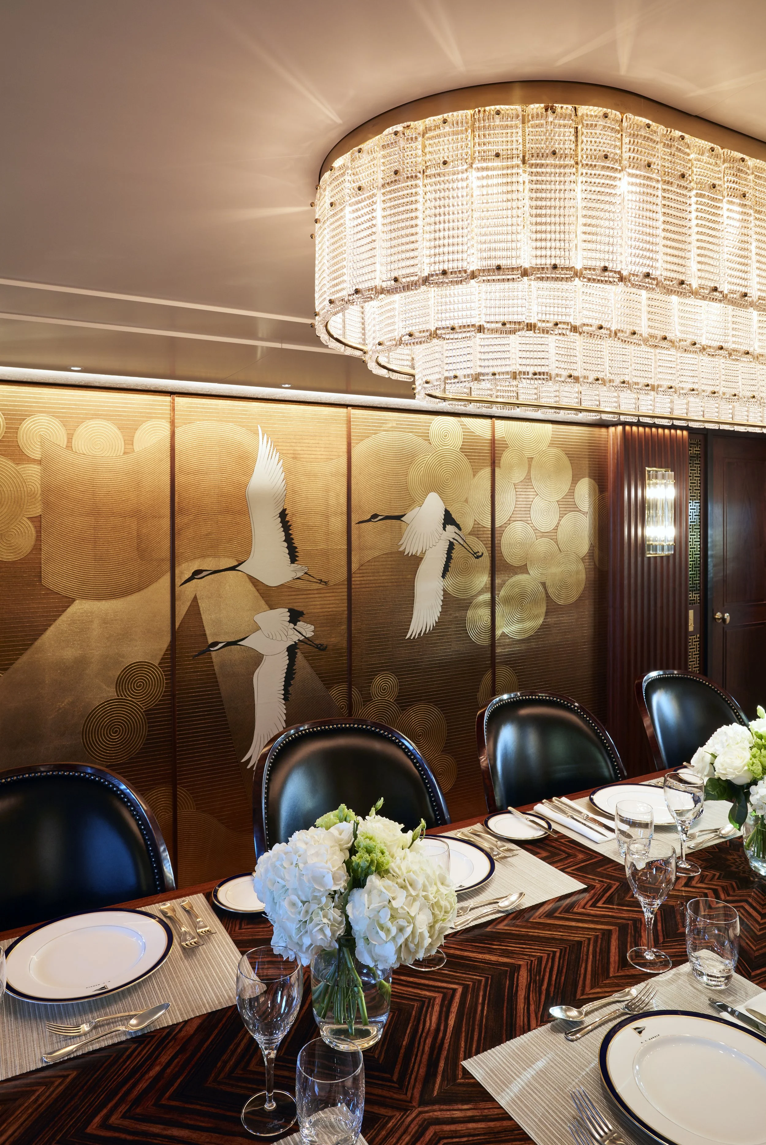 Elegant dining room with ornate chandelier, paneled wall art featuring cranes, polished wooden table set with white plates, glasses, cutlery, and floral centerpieces of white flowers.