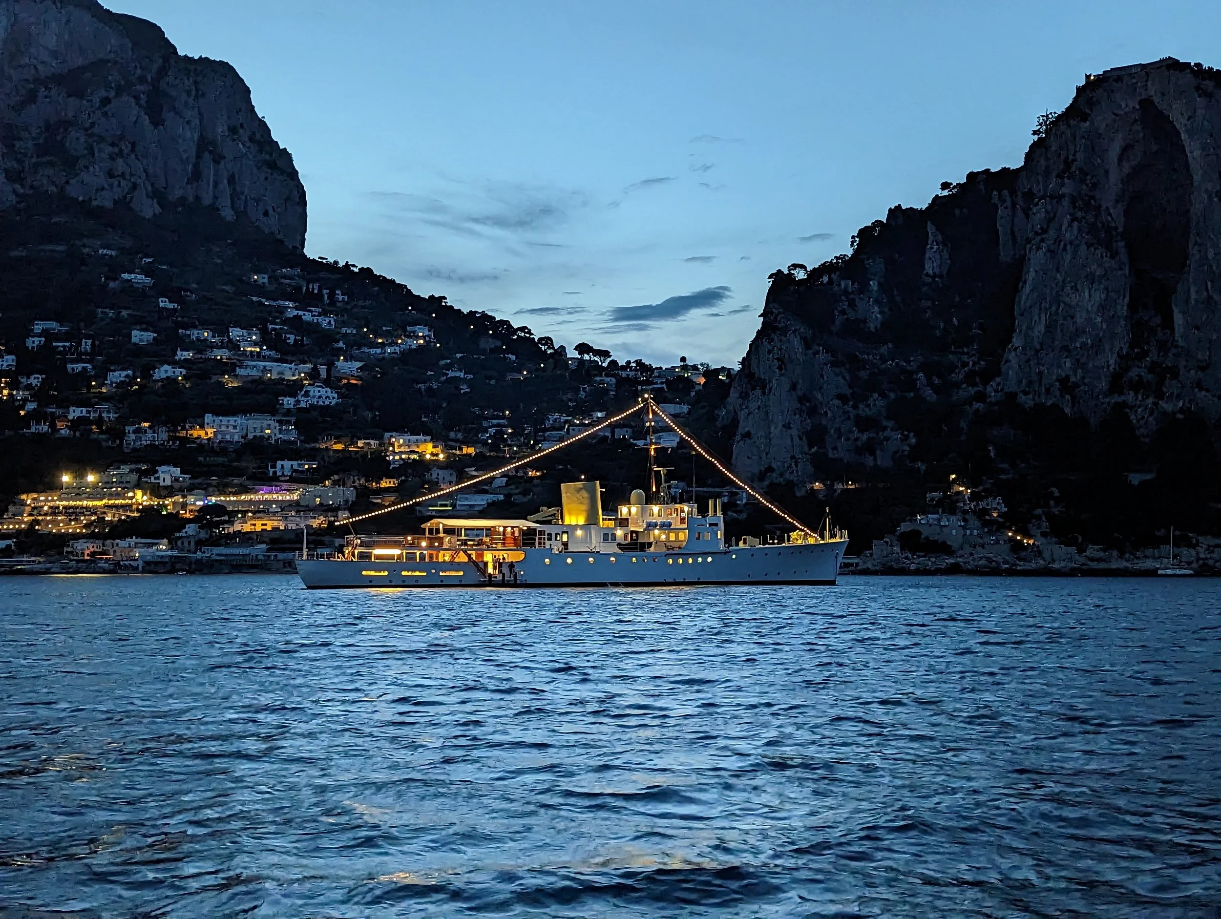 A yacht illuminated at night on water, surrounded by mountainous terrain and city lights in the background.