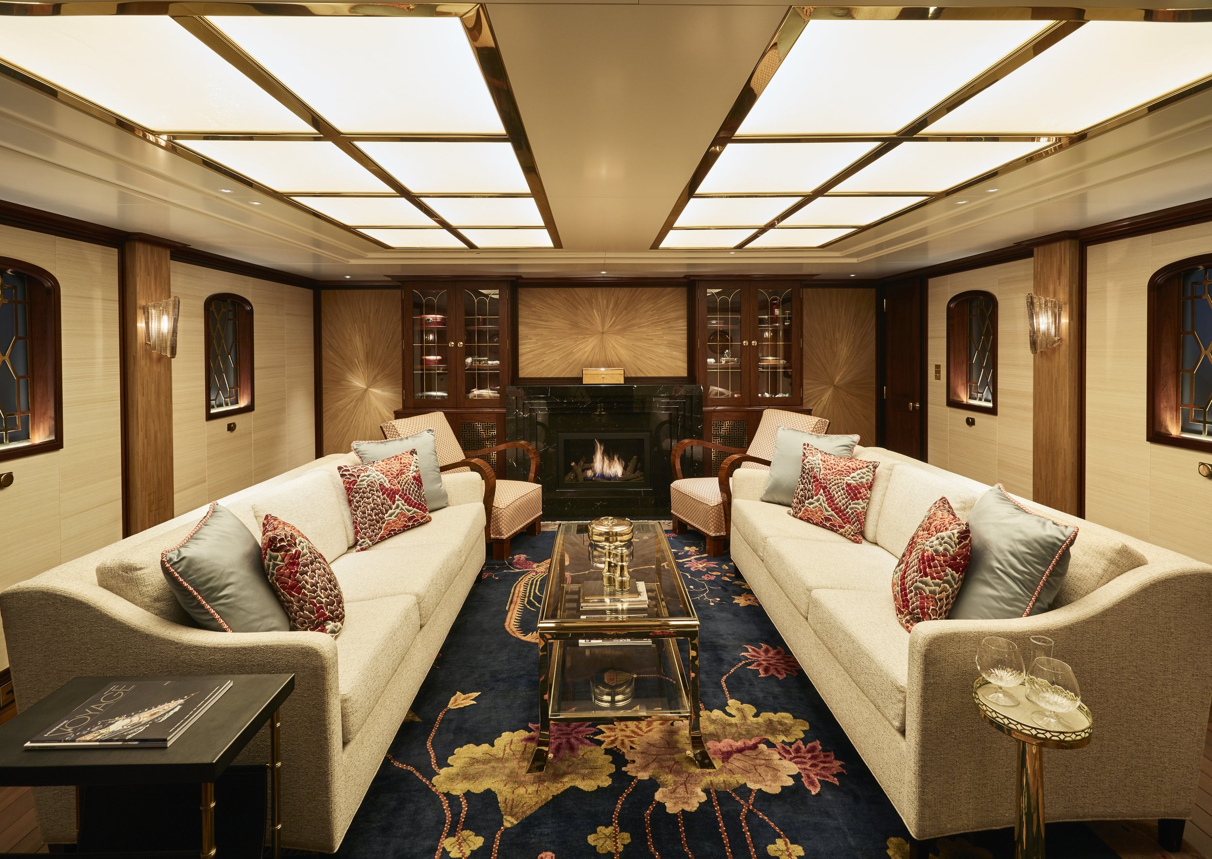 Luxurious yacht lounge with beige sofas, red decorative pillows, glass coffee table, floral carpet, and a fireplace flanked by shelves.