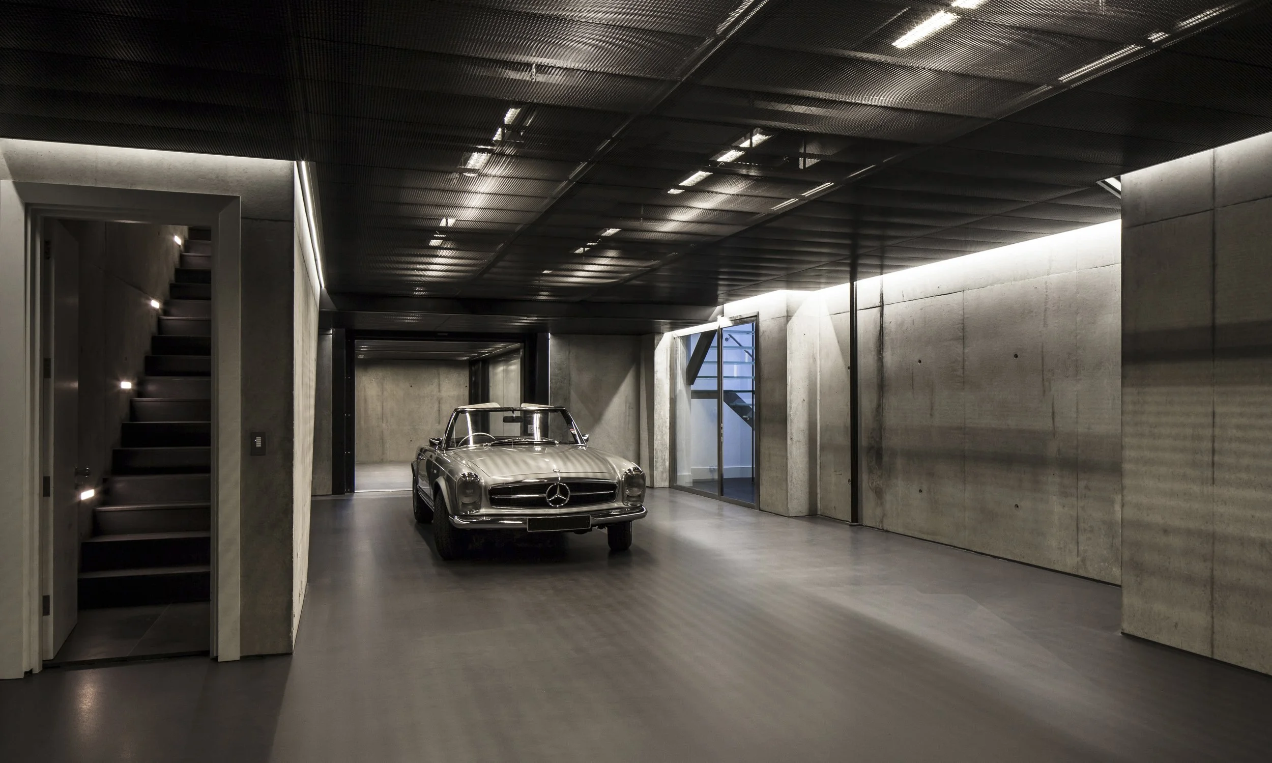 A modern, minimalist garage with a vintage silver Mercedes-Benz parked inside. The garage has concrete walls, a dark wood ceiling, and subtle lighting along the staircase and ceiling.