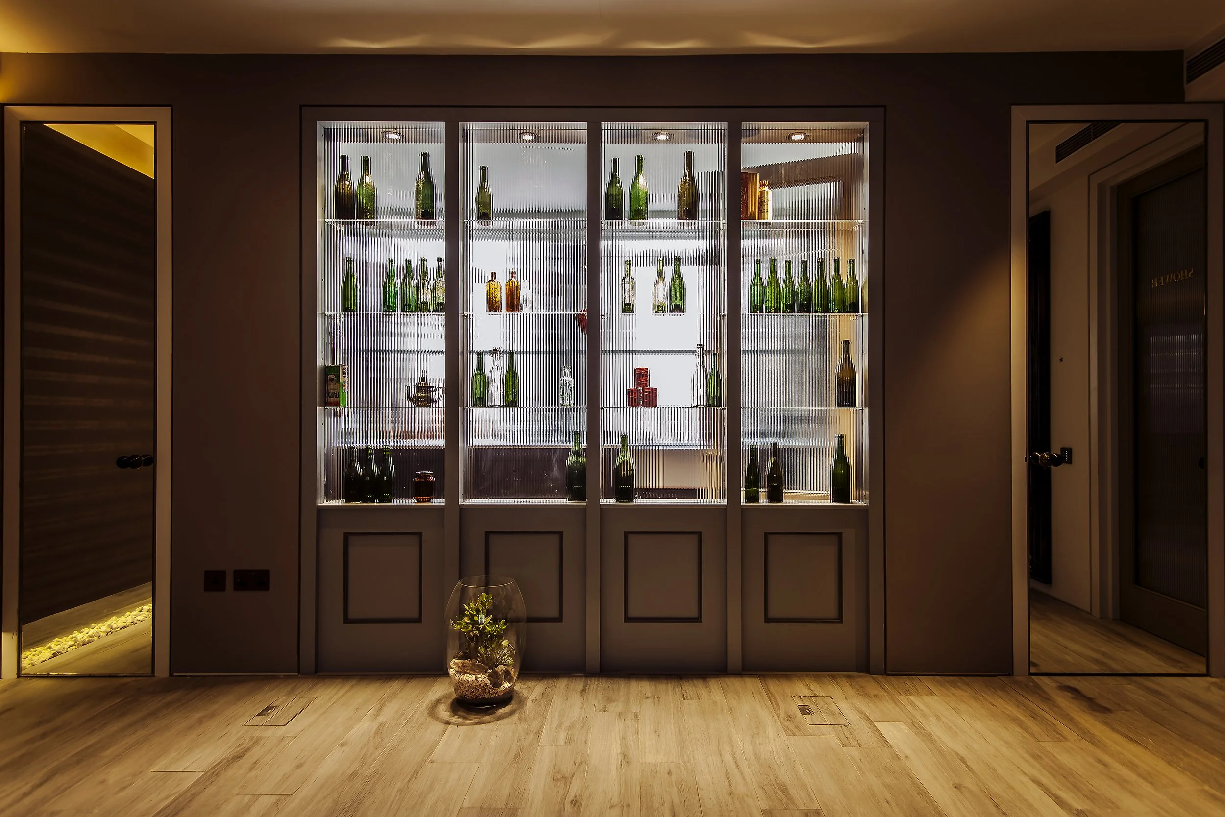 Glass cabinet with shelves displaying green and amber bottles, decorated with small ornaments, in a room with wooden floor and neutral walls.