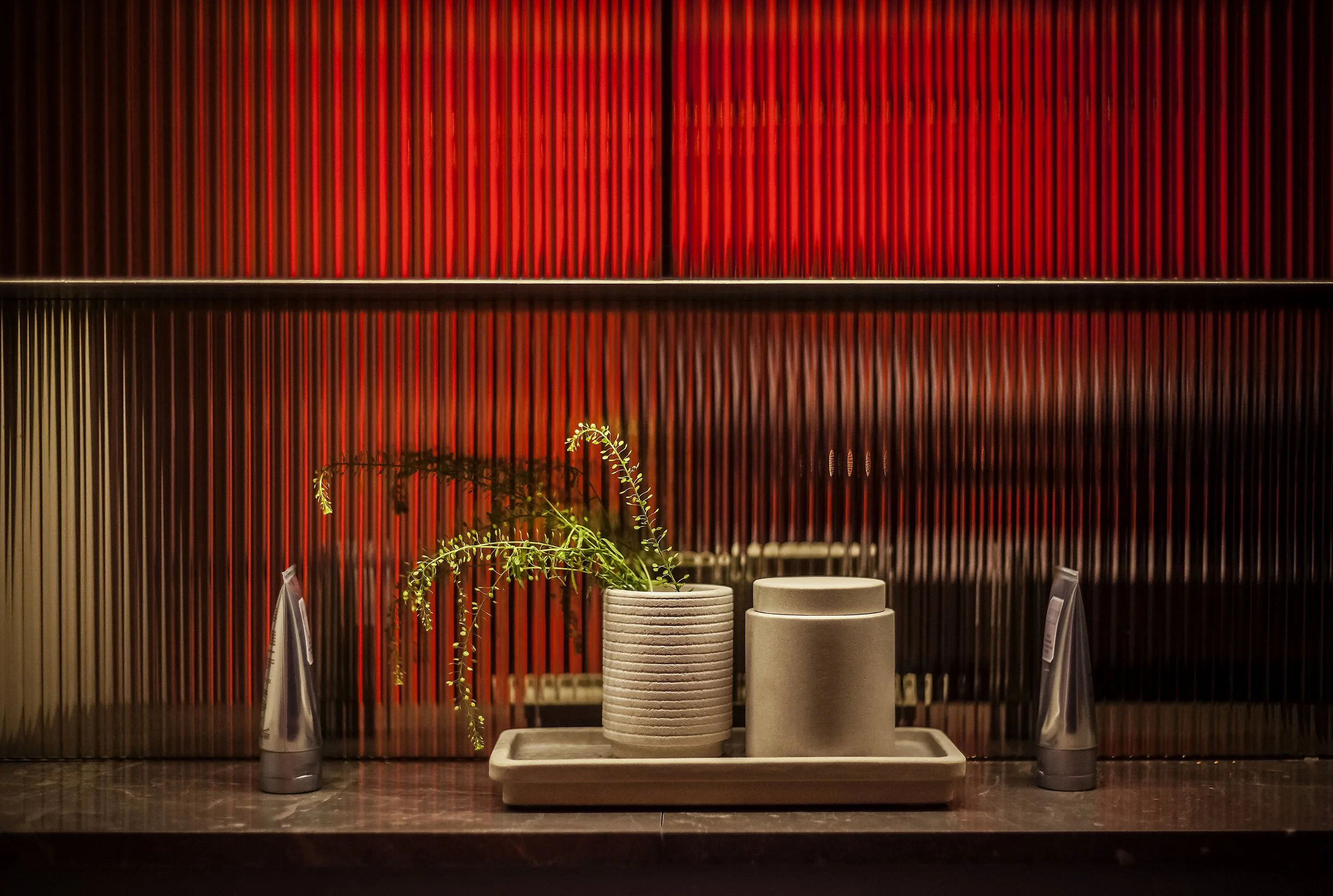 A decorative arrangement with two beige containers, a tray, a green plant, and two silver cylindrical bottles in front of a red ribbed wall with a reflective surface.